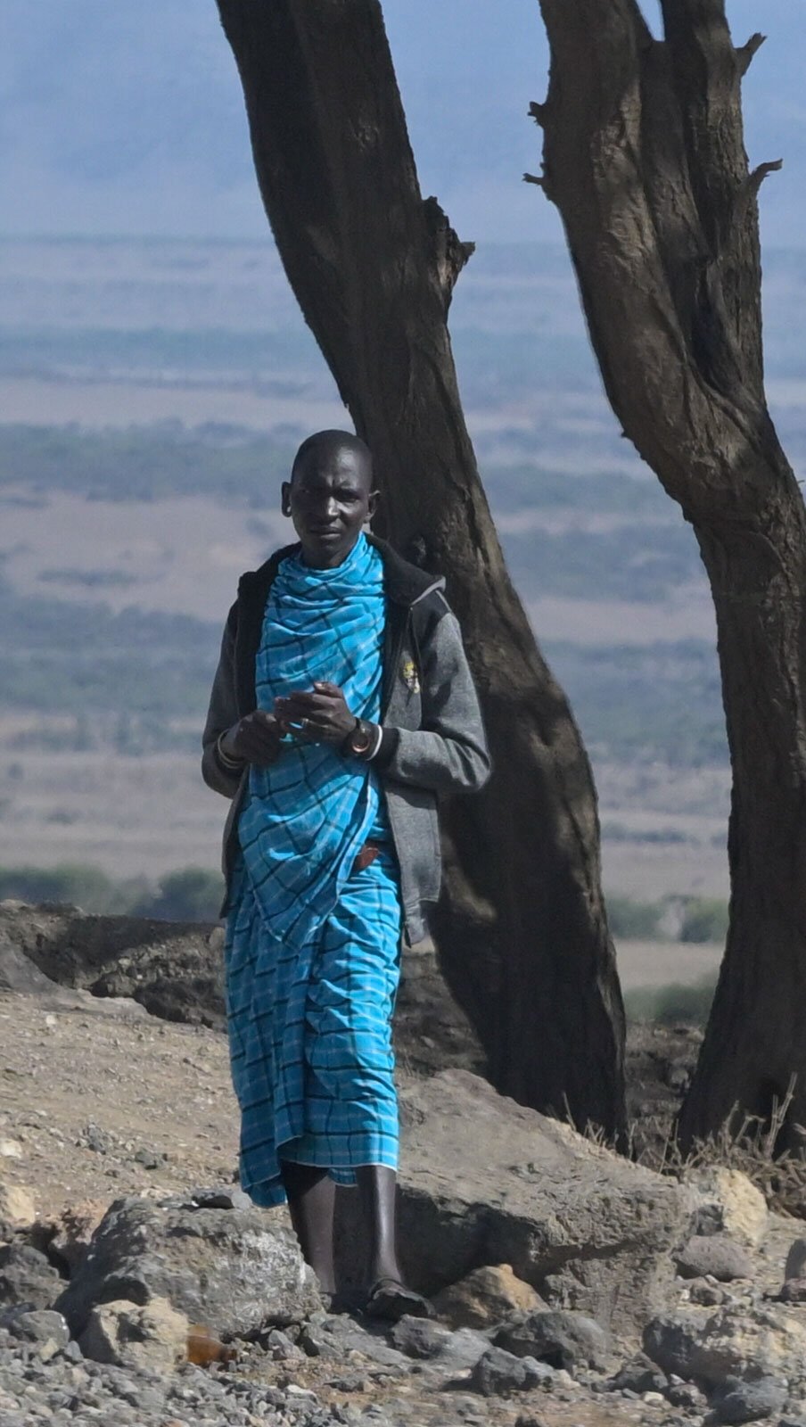 A person standing on rocky ground, wearing a blue patterned traditional garment and a gray jacket, with a blue scarf around the neck, near two large trees with a landscape in the background.