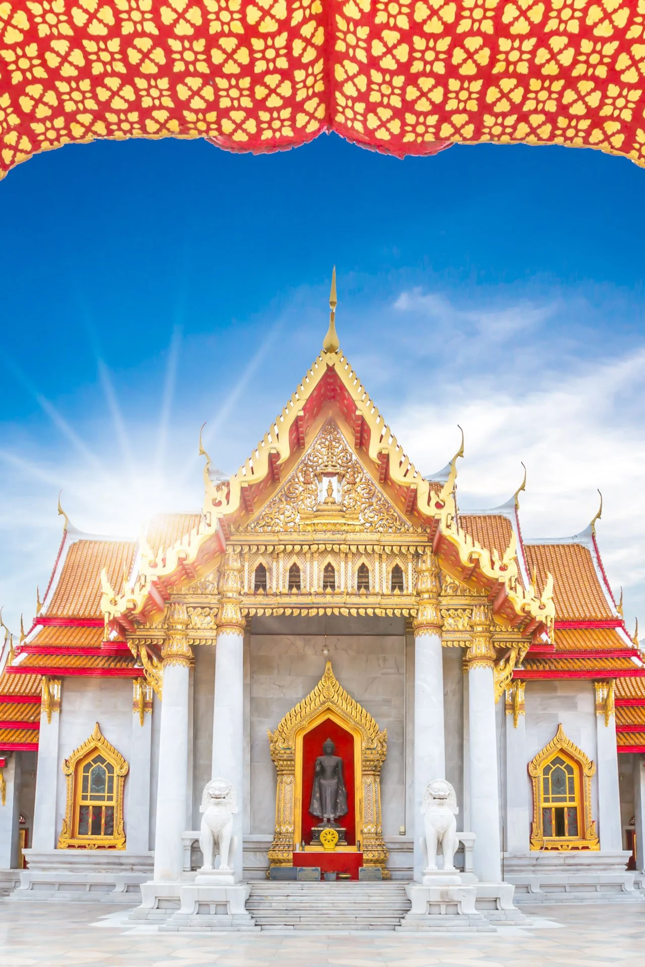 Front view of a Thai temple with ornate golden decorations, red accents, and statues of lions, under a blue sky with sunlight in the background.