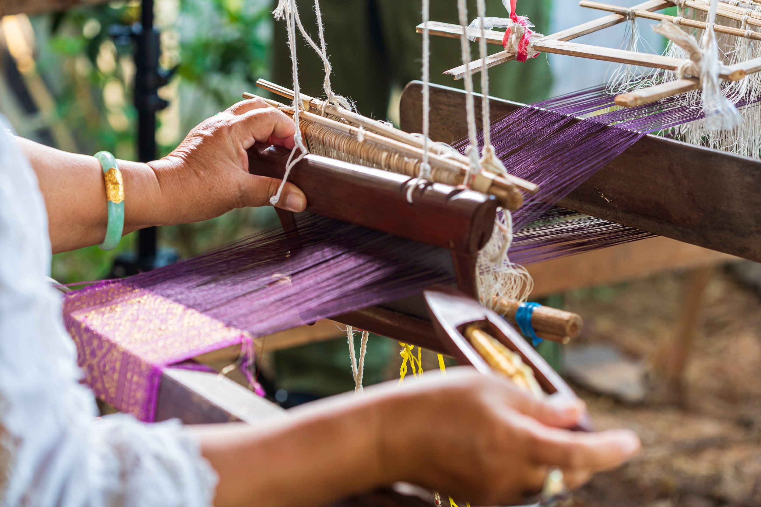 A person weaving on a traditional handloom with purple thread, involving wooden and bamboo parts, outdoors.