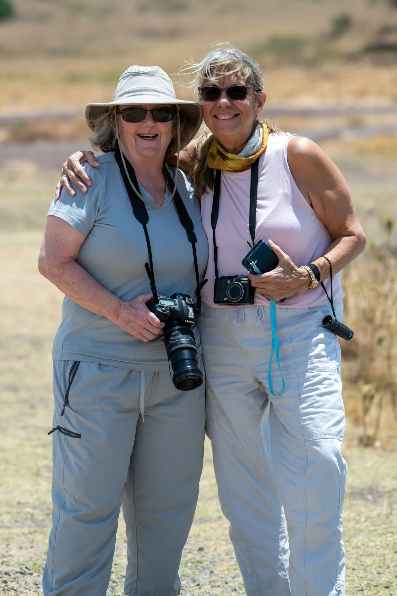 Two elderly women standing outdoors, smiling with their arms around each other, wearing sunglasses and casual hiking clothes, each holding a camera.