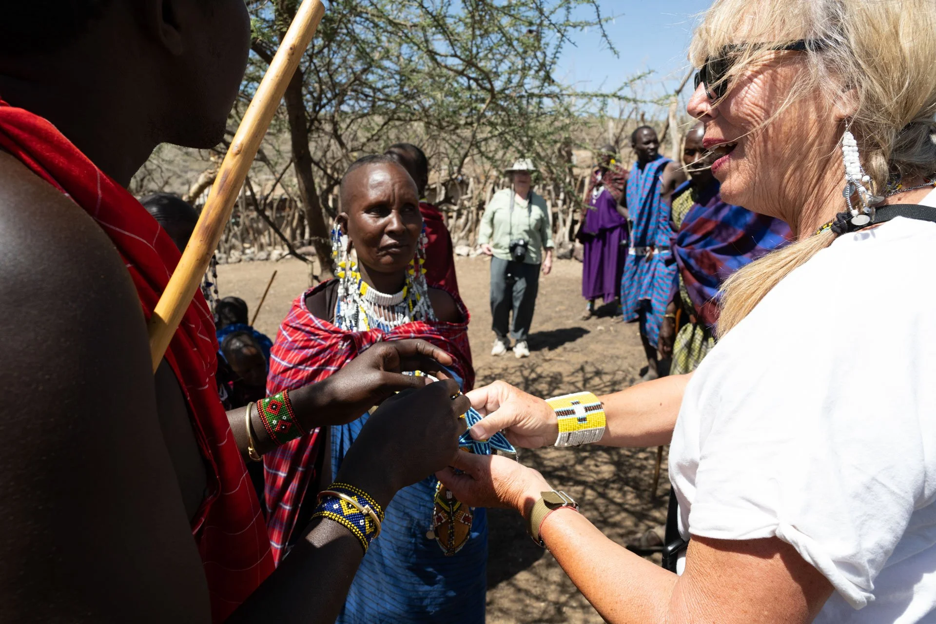 A woman with light skin and blonde hair wearing a white shirt and beaded jewelry hands a beaded bracelet to an indigenous woman with dark skin, wearing traditional Maasai clothing and jewelry, in a dry outdoor setting with trees and other people in t
