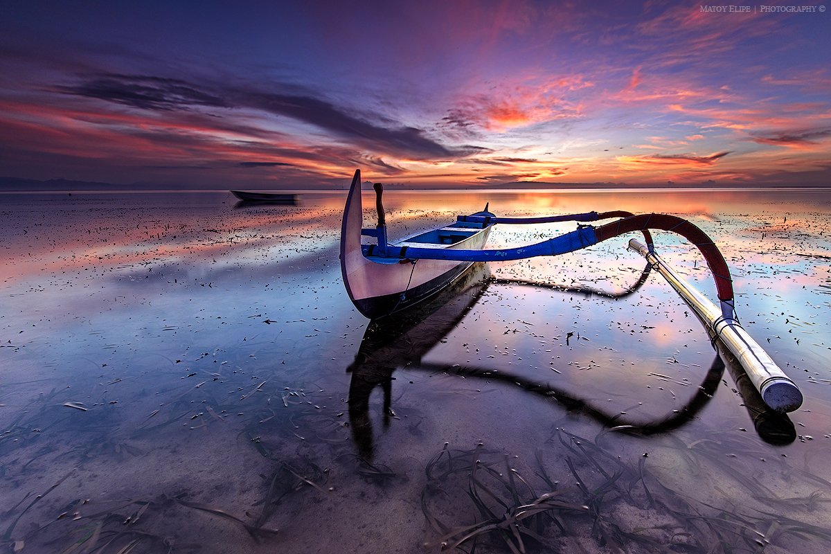 A boat with an outboard motor resting on the shore during sunset, with a colorful sky and calm water reflecting the sunset colors.