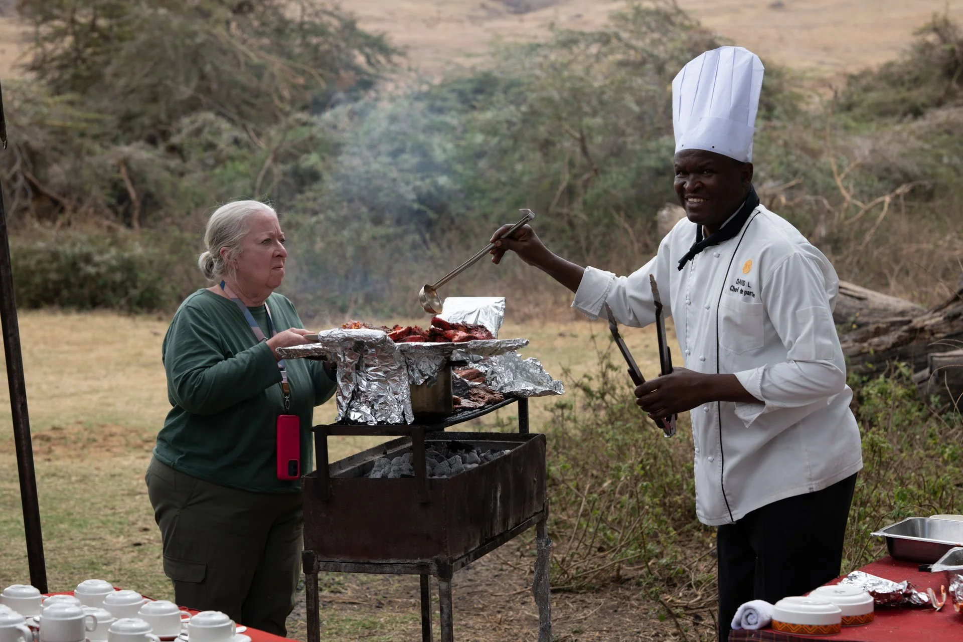 A chef serving grilled meat to a woman outdoors at a rustic barbecue or outdoor cooking event in a natural setting with trees and grass.