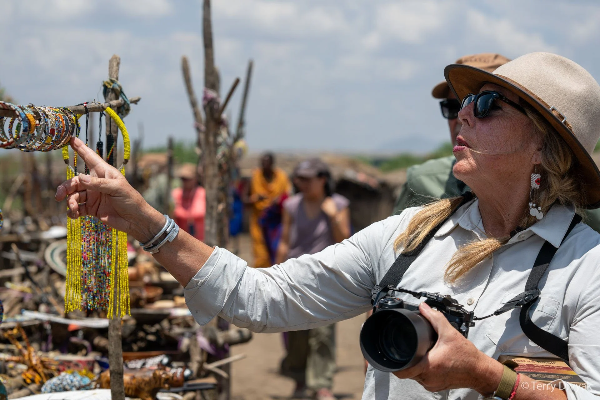 A woman with sunglasses and earrings points at colorful bracelets on a display stand, with a camera hanging around her neck, in a Maasai marketplace setting.