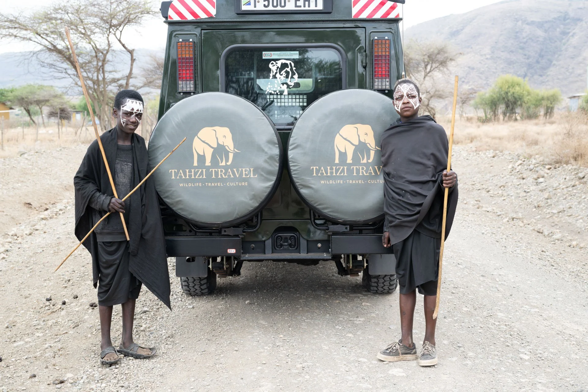 Two Maasai Boys dressed in traditional Maasai clothing with face paint, standing outdoors in front of a safari vehicle with large spare tires labeled 'Tahzi Travel.' The landscape is dry with sparse trees and hills in the background.