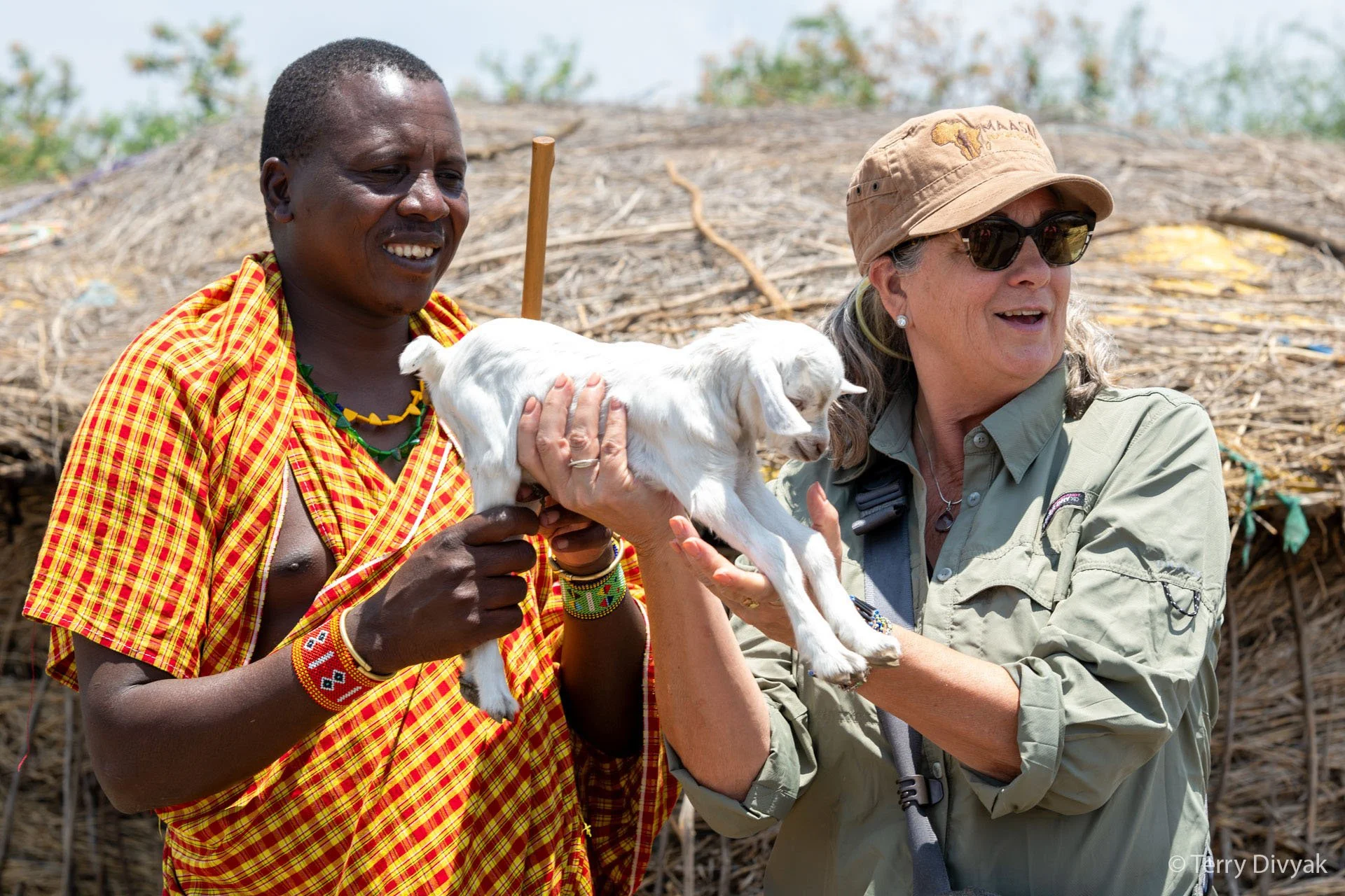 Two women, one indigenous and the other wearing a hat and sunglasses, are holding a small white goat outdoors. The woman with the hat is smiling and wearing a khaki shirt, while the other woman is dressed in traditional Maasai attire with colorful be