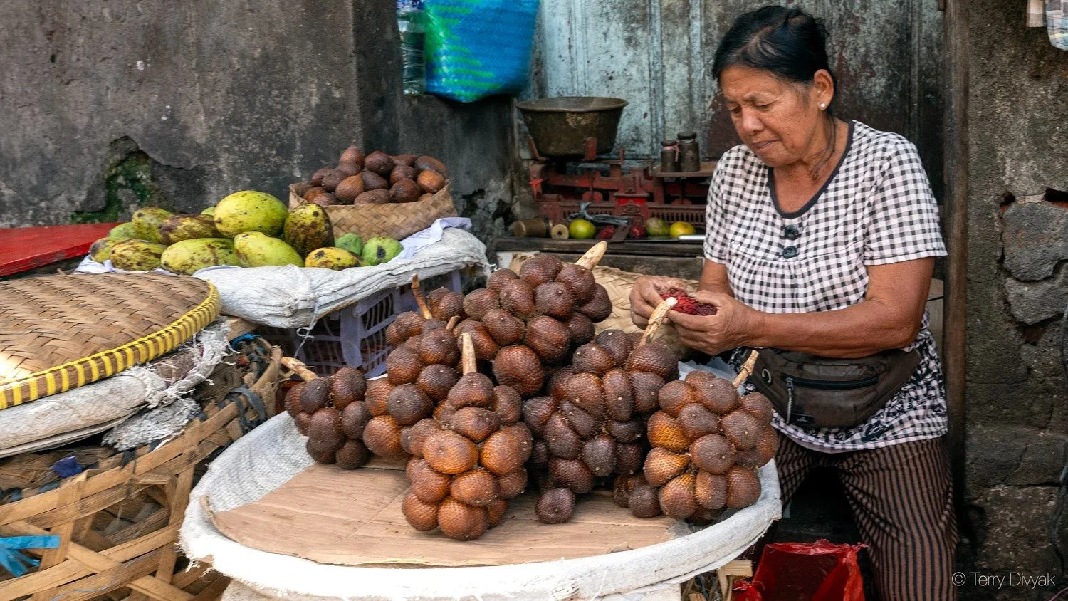 An older woman standing at a market stall sorting and peeling on-looking at clusters of salacca, a type of tropical fruit with scaly brown skin and a reddish interior, with other fruits like bananas and avocados on display.