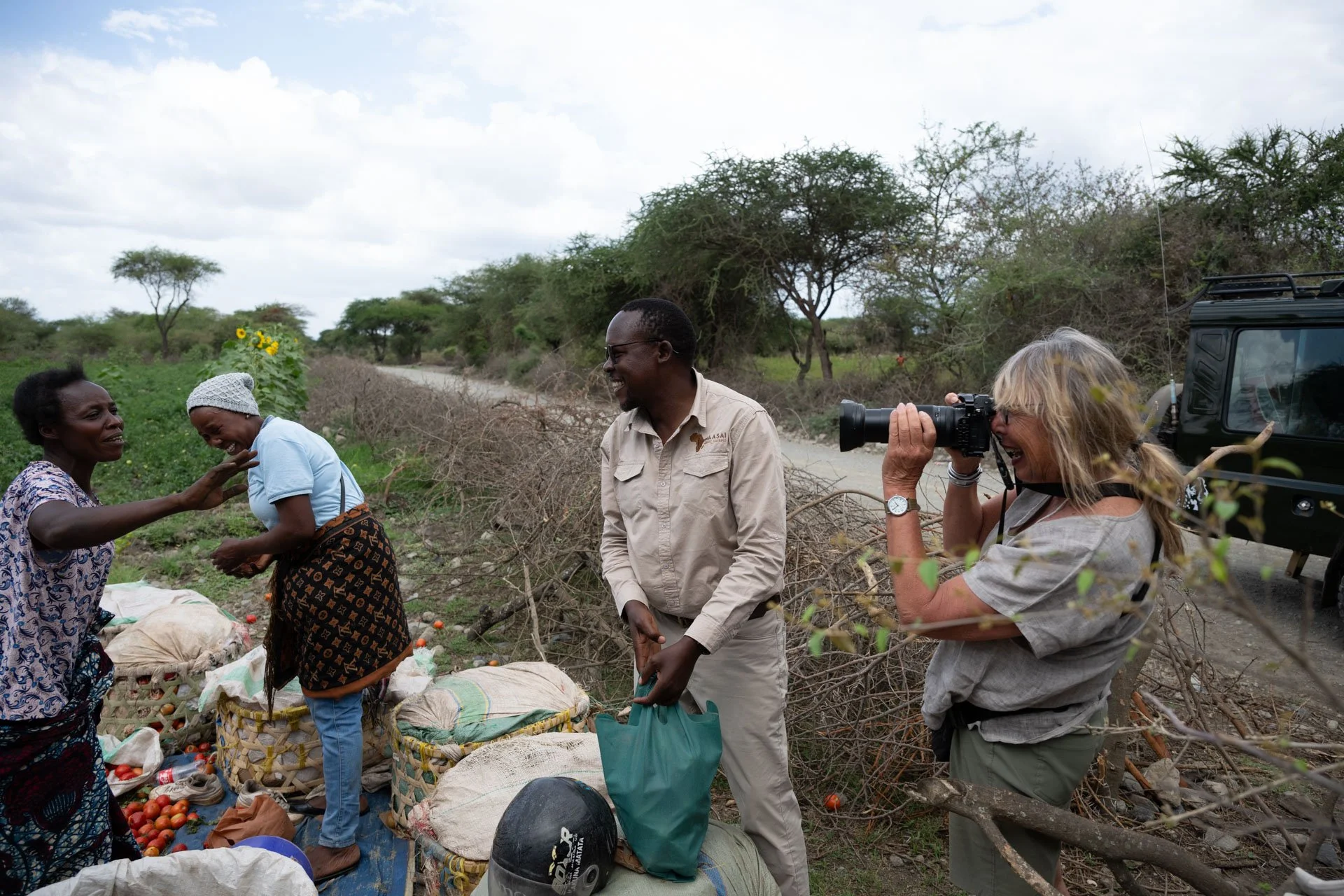A group of people, including a woman with a camera, are outdoors on a rural dirt path with trees and bushes in the background. Two women are engaged in conversation near a pile of tomatoes and sacks.
