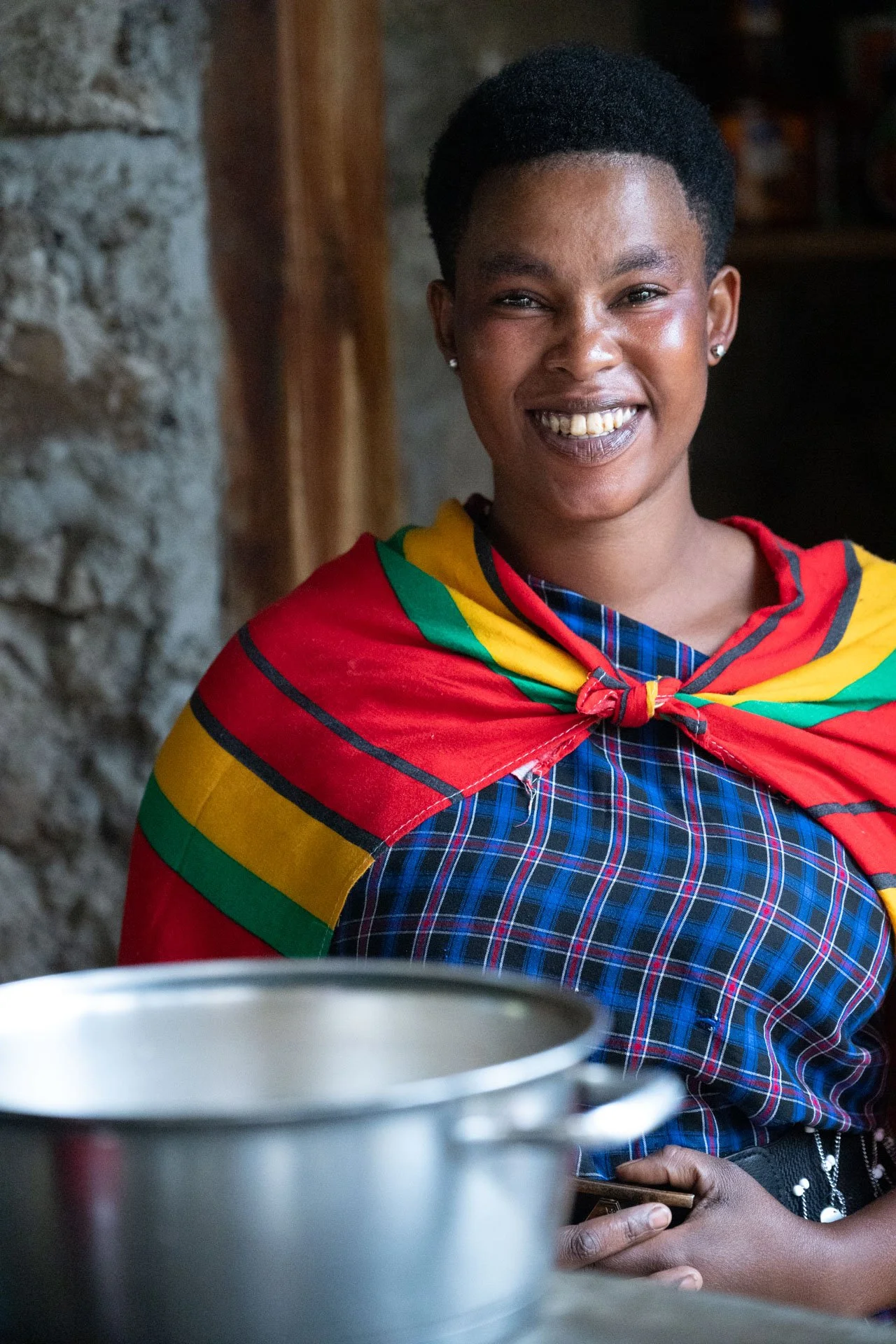 A smiling woman wearing a colorful striped sash over a blue plaid dress, standing indoors with a stone and wood background, near a metal pot.