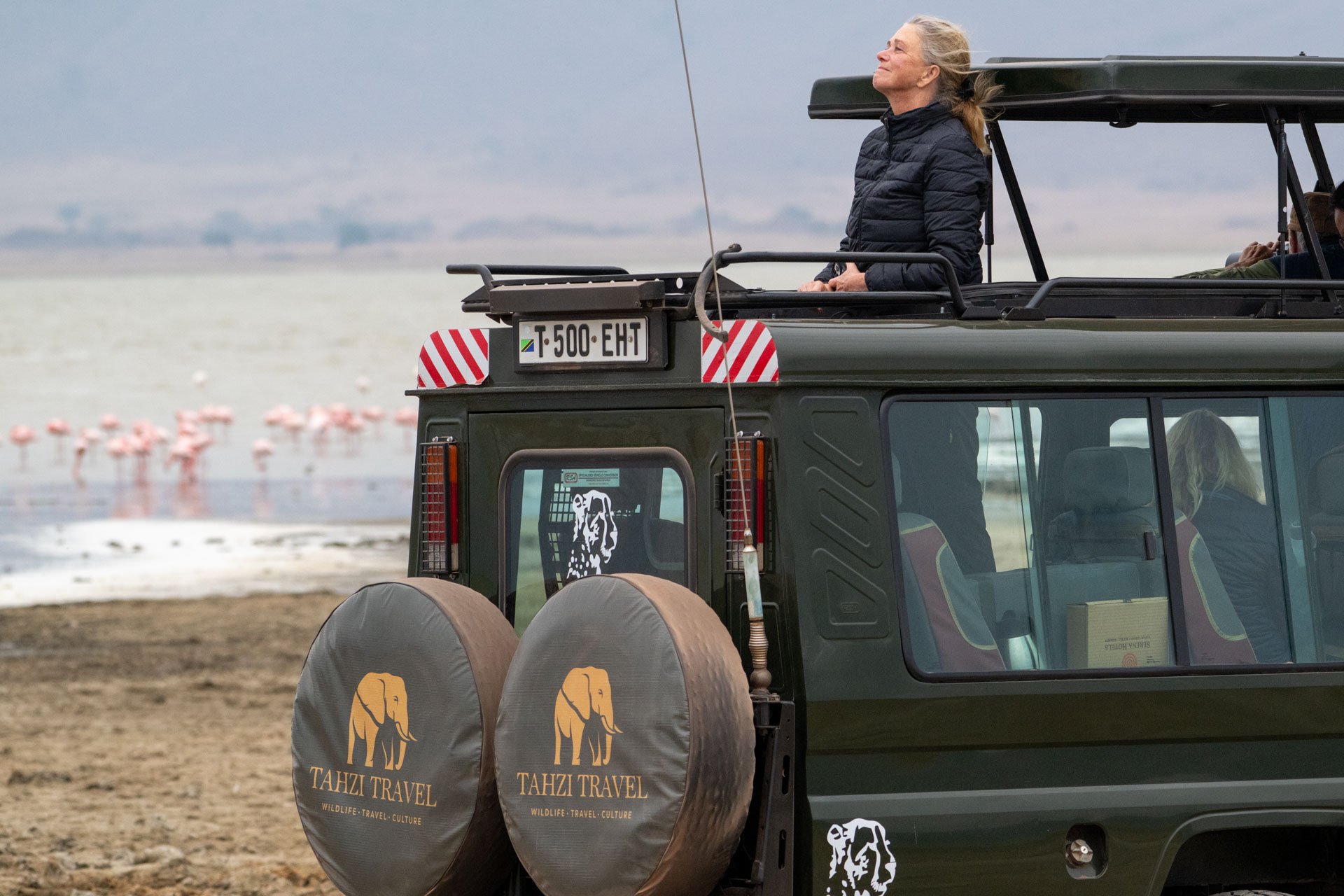 A woman with gray hair wearing a black jacket stands on a safari vehicle with a roof, enjoying the view of a lake with pink flamingos in the background.