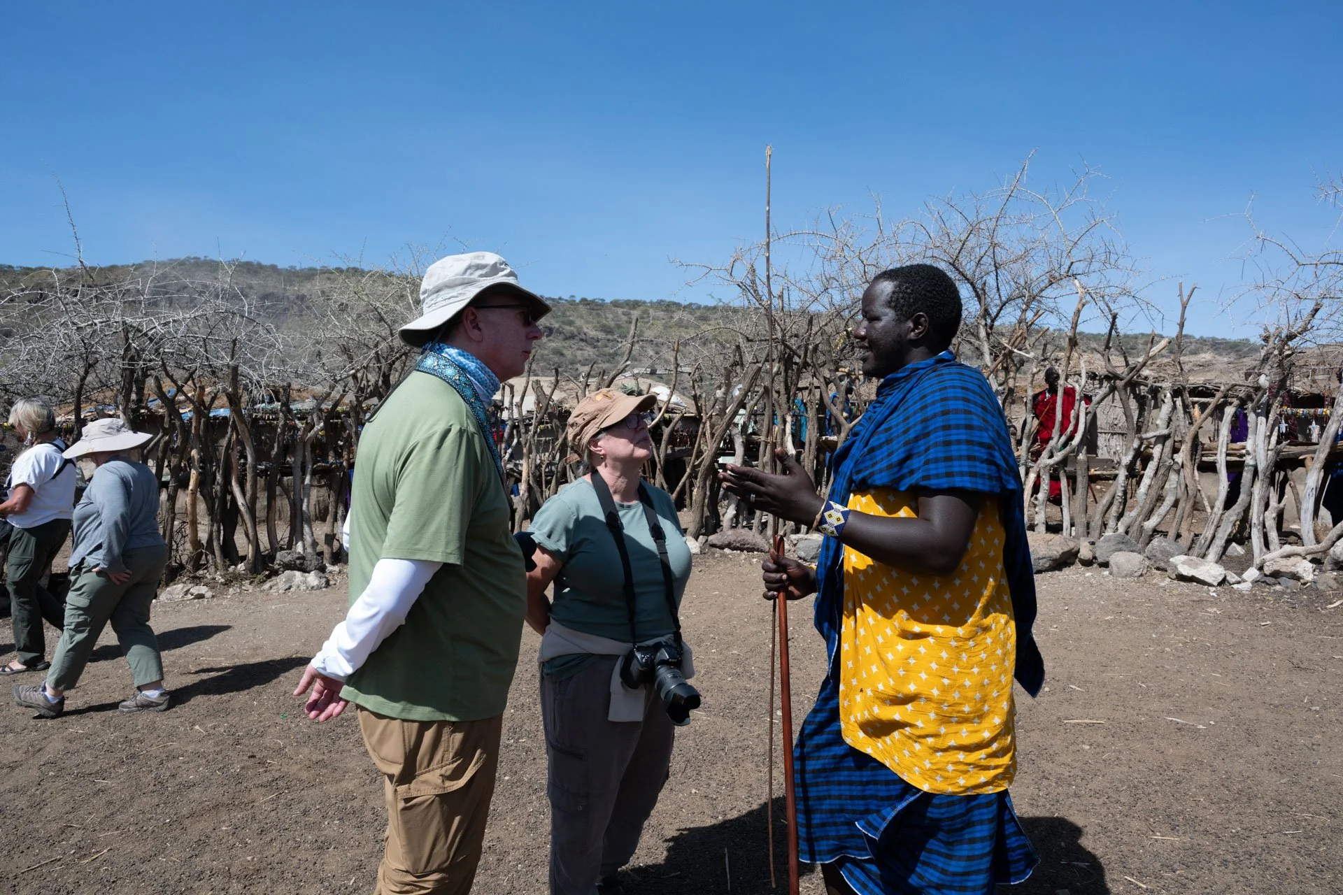 Three people are engaged in a conversation outdoors in a dry, rural area with leafless trees. Two tourists with hats and cameras are speaking with a Maasai man dressed in traditional brightly colored clothing, holding a walking stick.