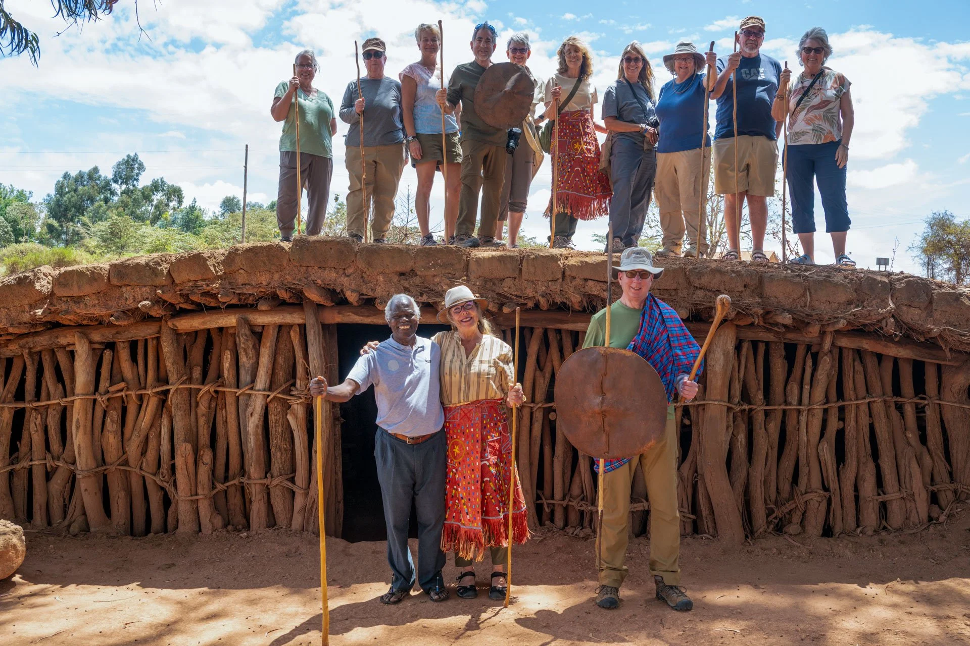 Group of people standing on top of and in front of a reconstructed ancient mud-brick shelter, posing for a photo outdoors under a partly cloudy sky.