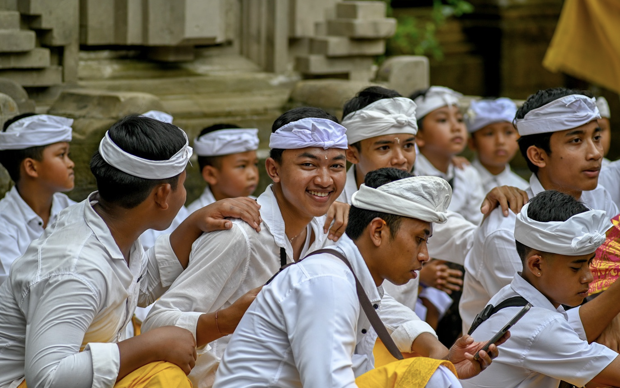 Group of young Balinese boys dressed in traditional white shirts and yellow sarongs, wearing white headbands, sitting outdoors on a staircase and engaging with each other.