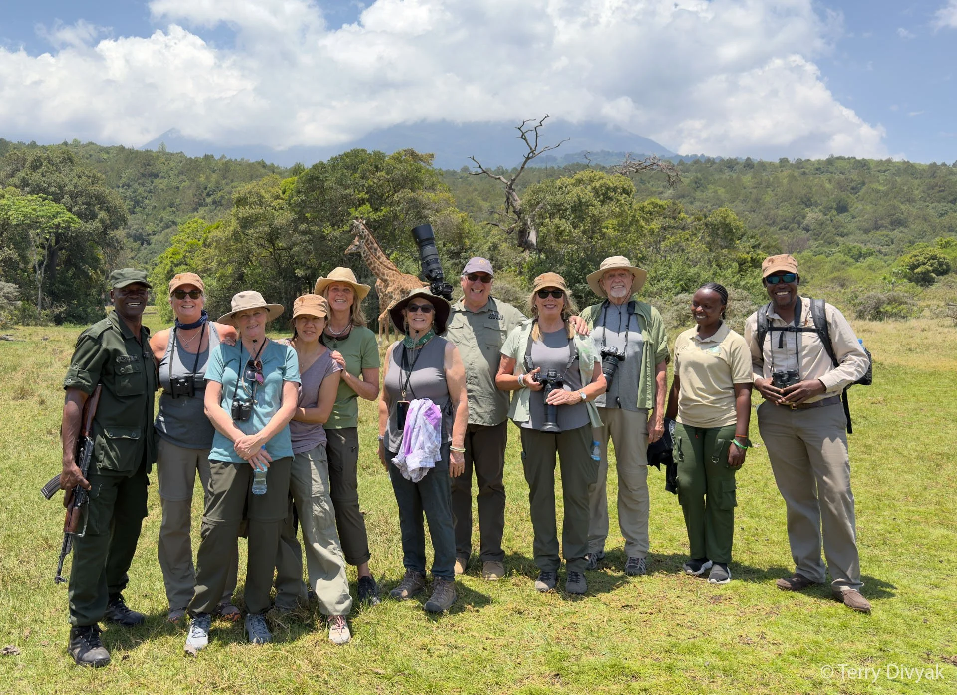 Group of people standing in a grassy field with a giraffe in the background, surrounded by trees and mountains.