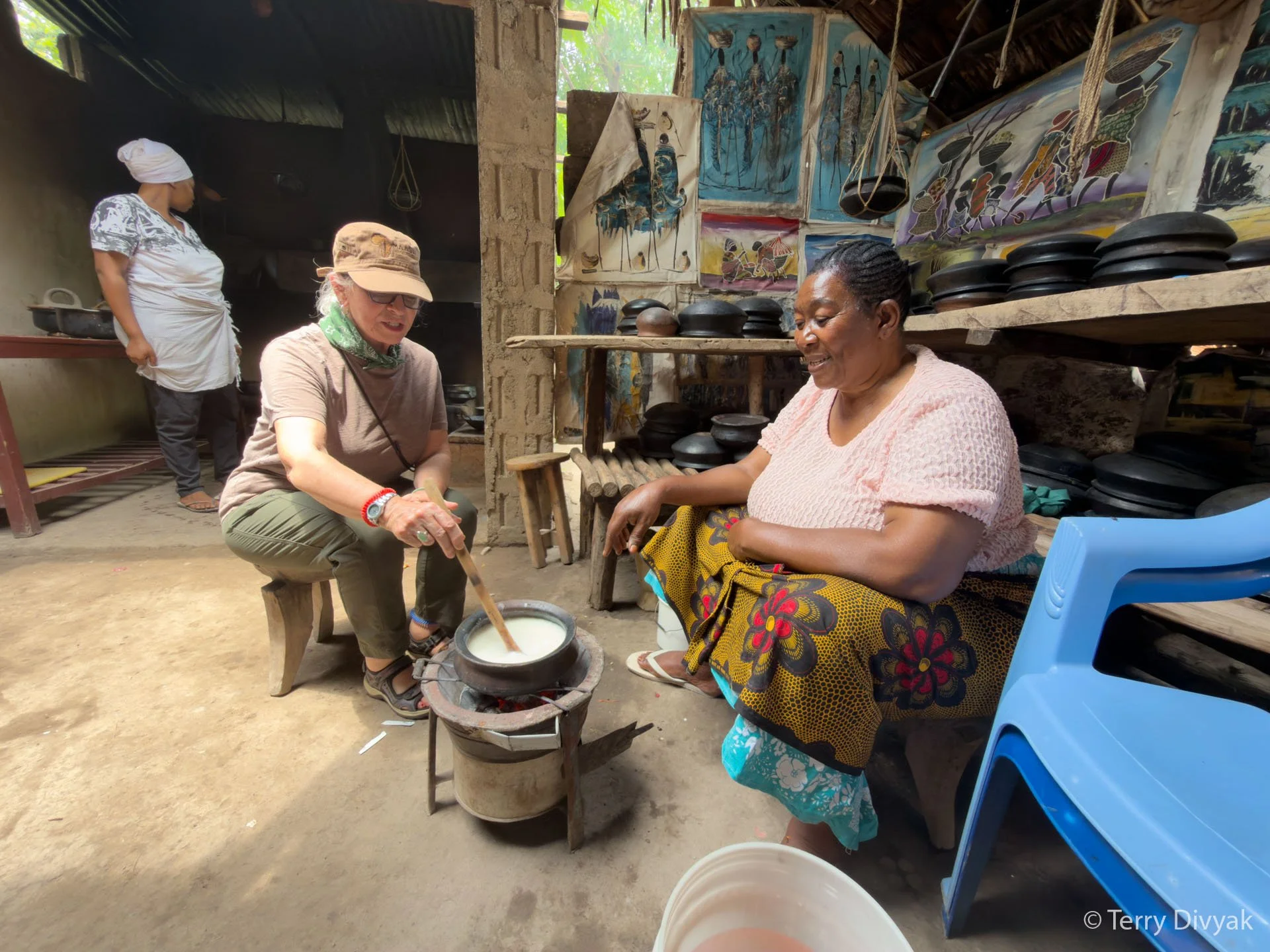 Two women are having a discussion, one is sitting on a plastic chair and the other on a small wooden stool. The woman sitting on the chair is wearing a pink top and a patterned skirt, and the woman on the stool is stirring a pot of white substance ov