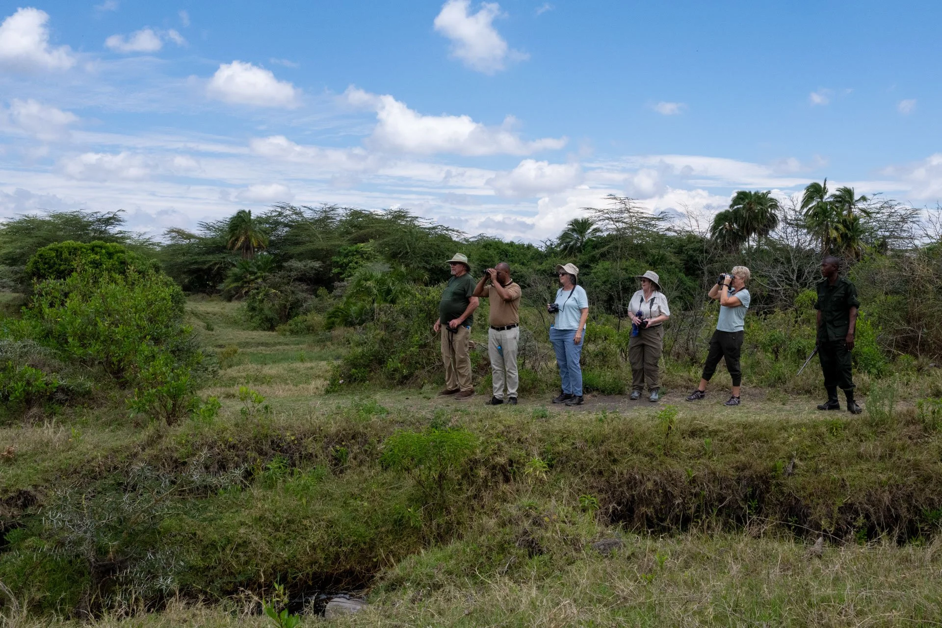 Group of six people standing outdoors in a grassy, wooded area on a walking safari tour with Tahzi Travel. Some with binoculars and cameras, observing wildlife or scenery under a partly cloudy sky.