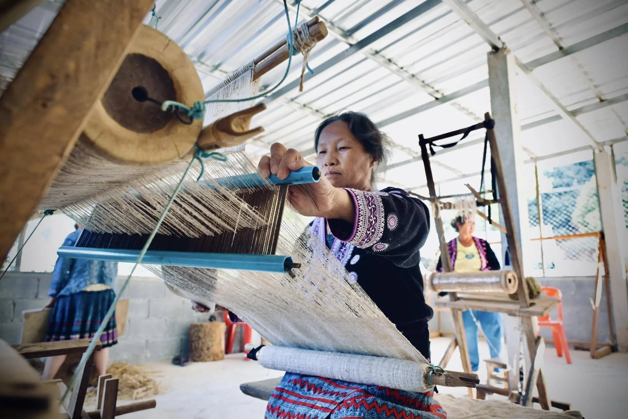 Woman weaving fabric on a loom inside a workshop with other people and weaving equipment.