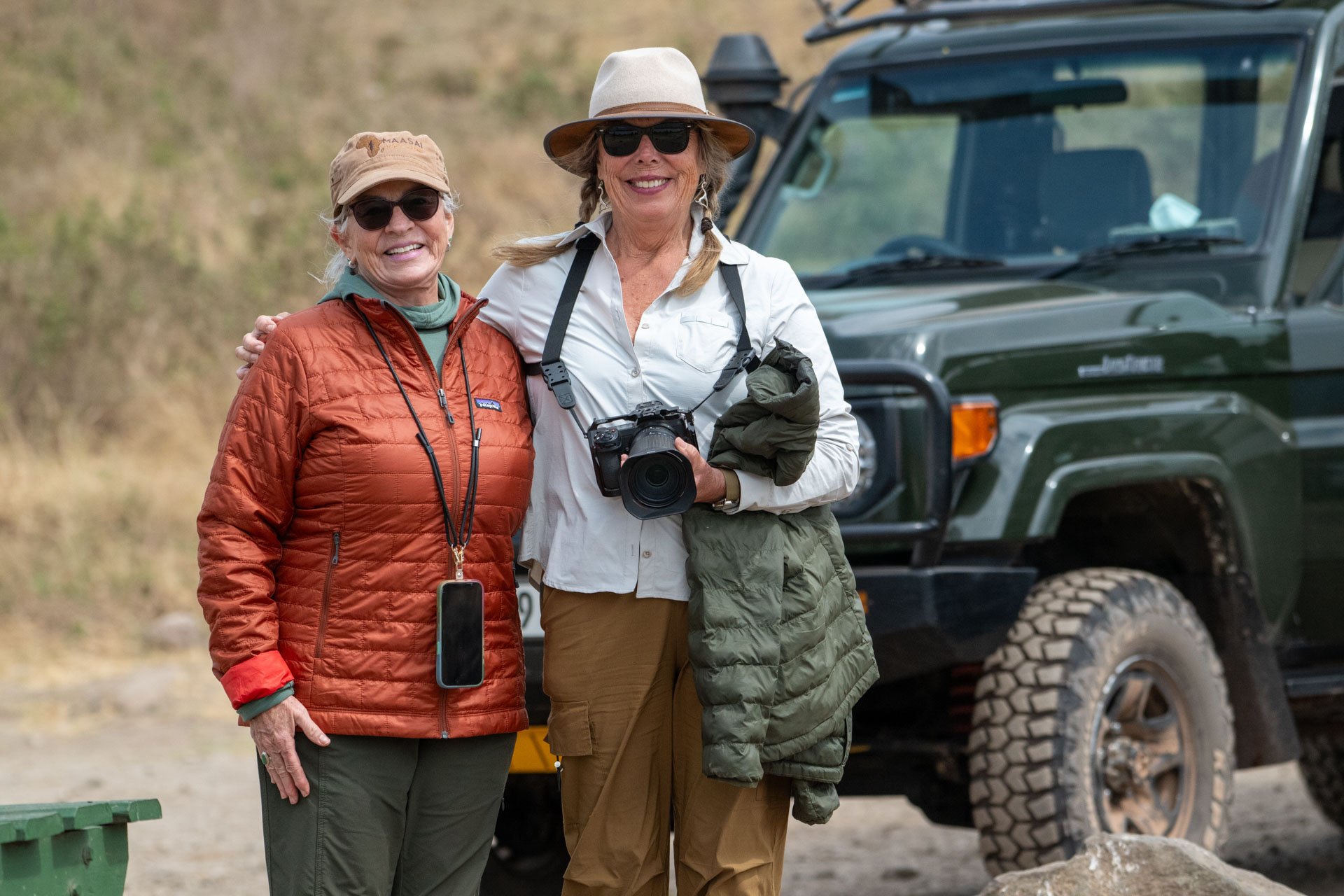 Two women smiling and posing outdoors in front of a green off-road vehicle. They are dressed in outdoor gear, with one woman wearing a red quilted jacket and the other wearing a tan hat and white shirt holding a camera.