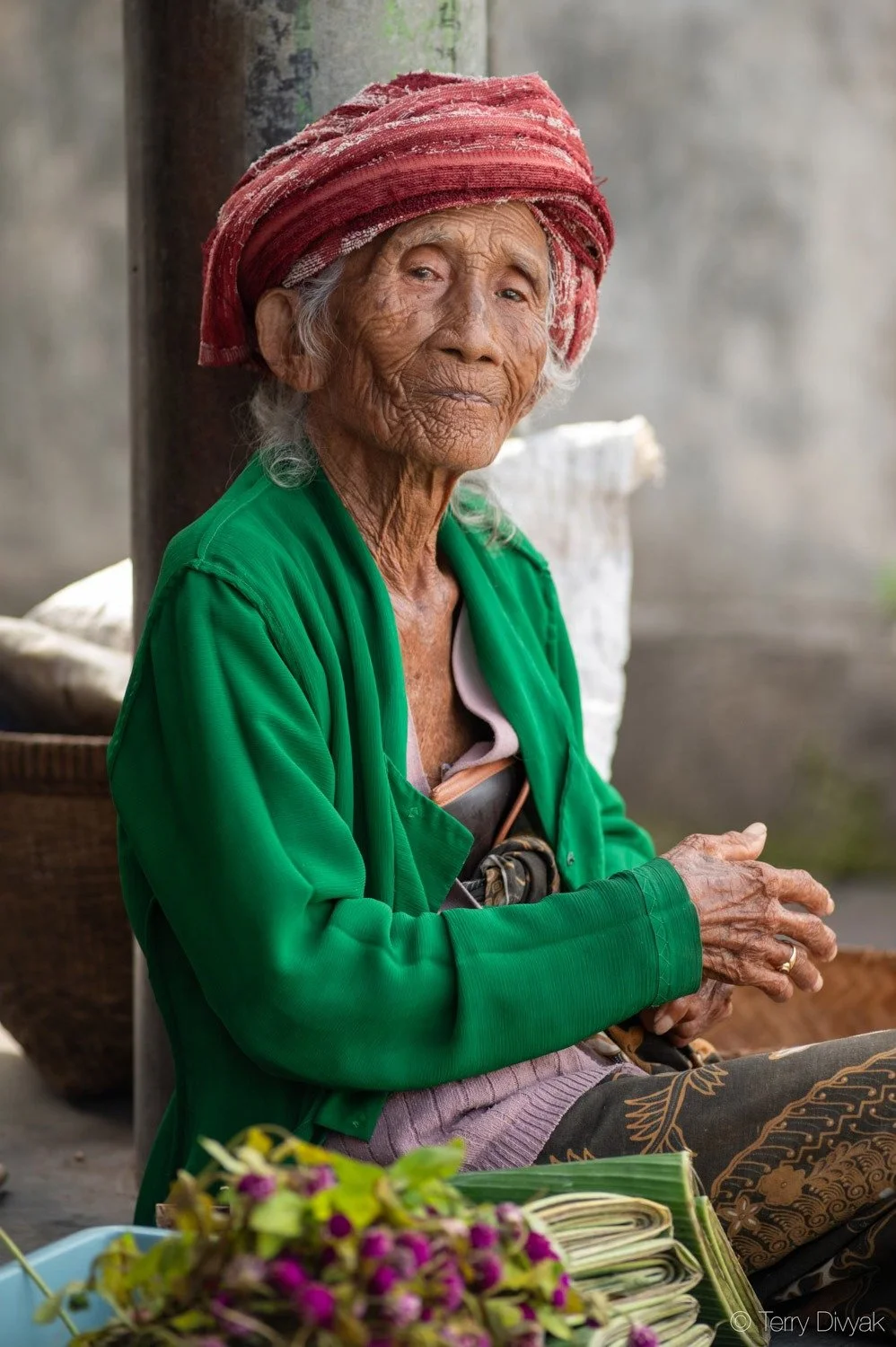 An elderly woman with a weathered face and gray hair, wearing a red headscarf and a green jacket, sitting outdoors with a thoughtful expression.