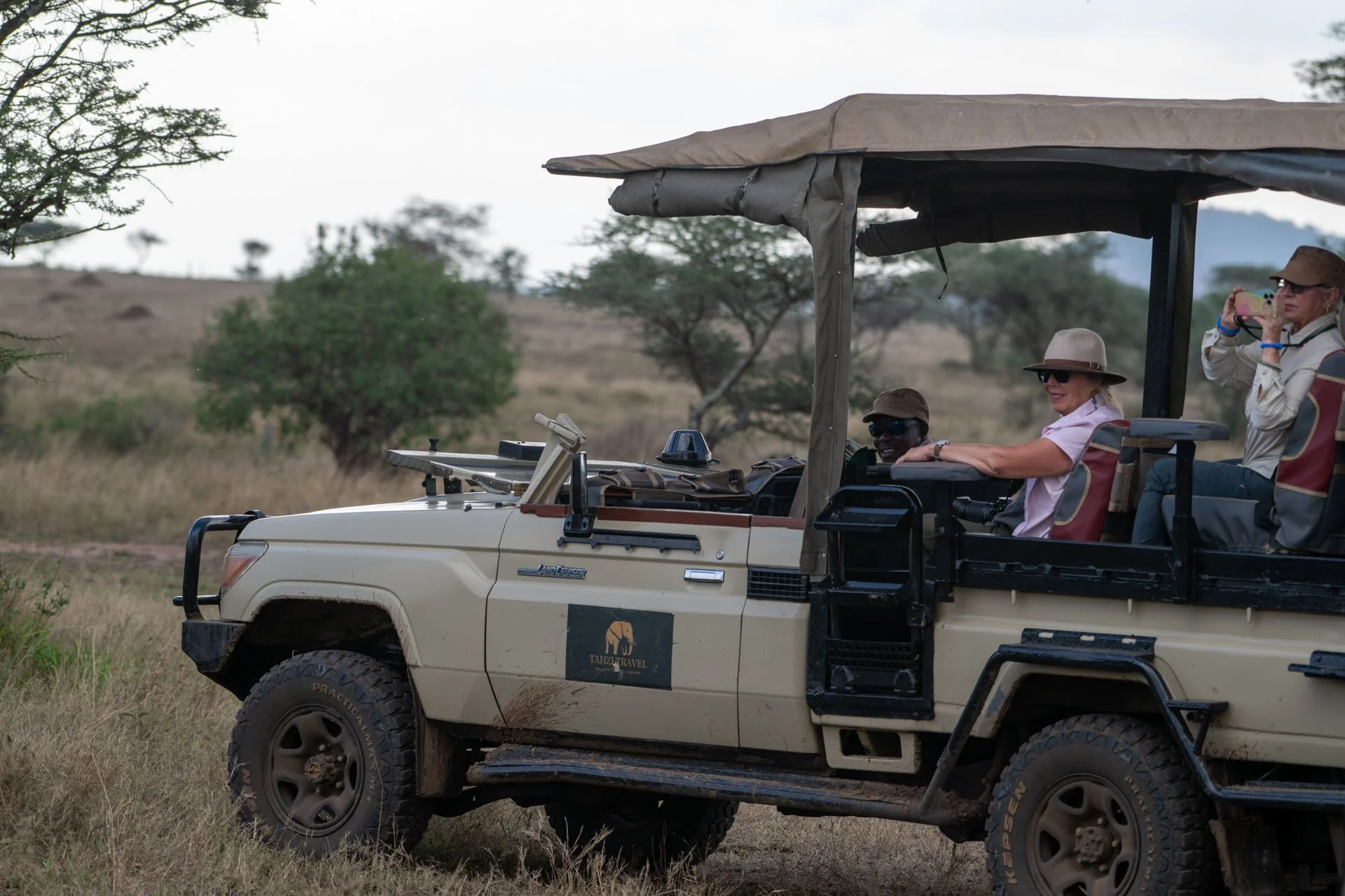 People on a safari vehicle with an open roof, taking photos in a savannah landscape with bushes and trees.