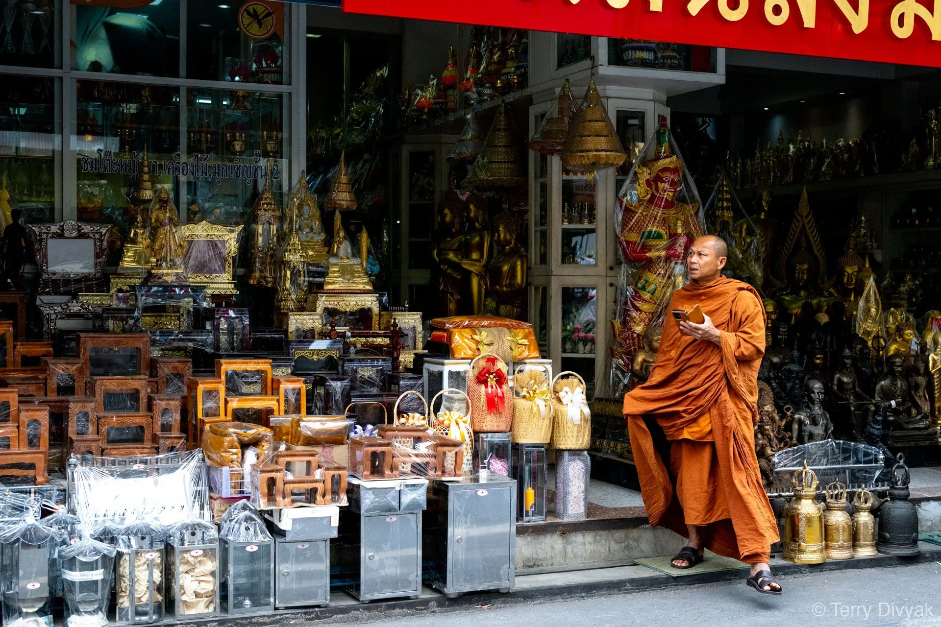 A Buddhist monk in orange robes walking past a shop selling religious statues and ritual items. The shop displays gold and bronze statues, wooden furniture, and decorated baskets outside.
