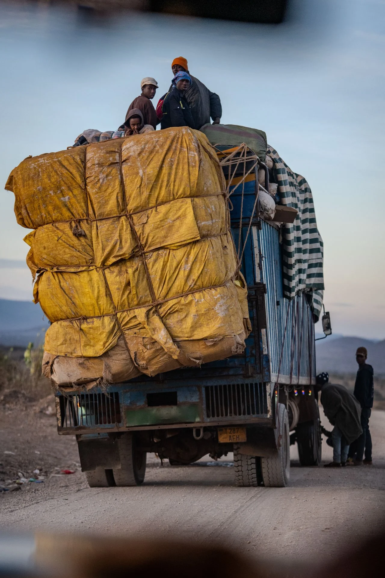 A group of people riding on top of a heavily loaded blue truck, with additional people standing next to it on the ground. The truck is on a dirt road with mountains in the background, during dusk or early evening.
