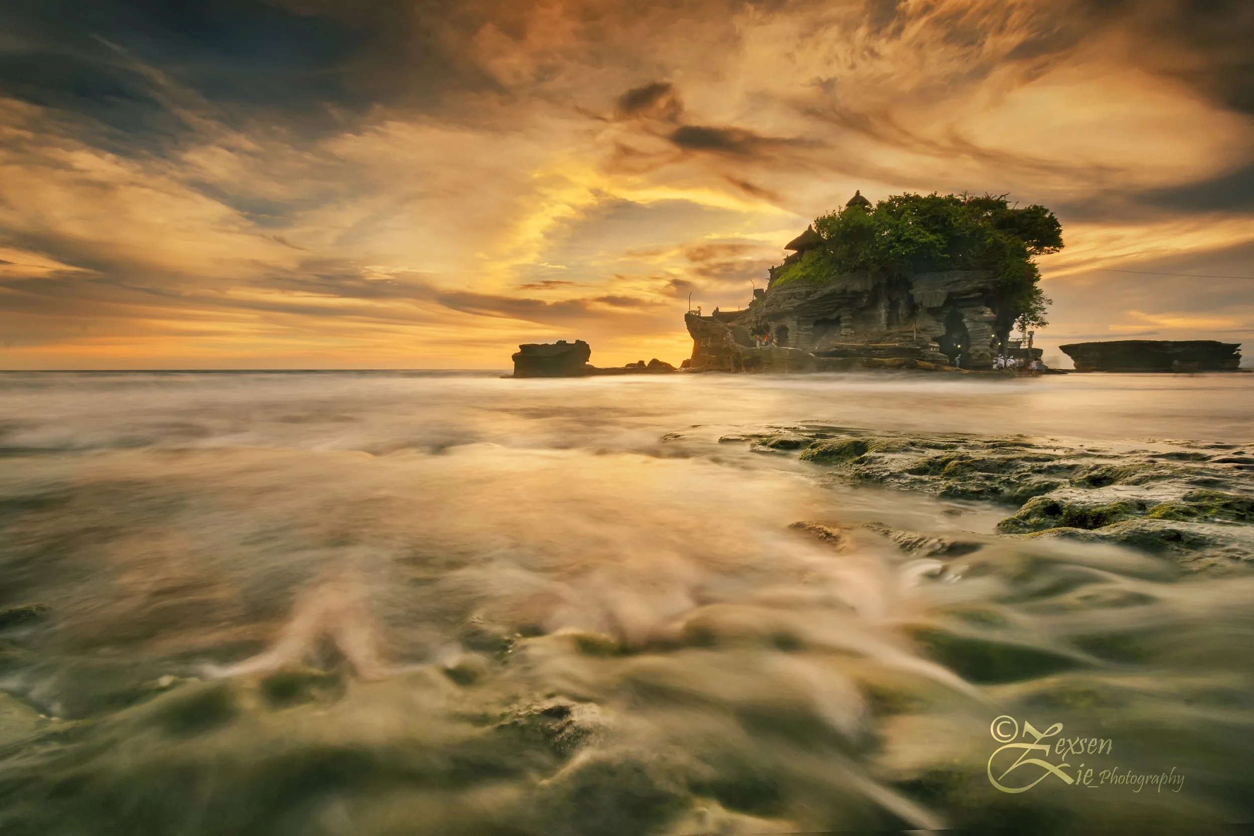 Sunset over rocky shoreline with a building on a small island covered in green trees, calm waves in the foreground, and a colorful sky with clouds.