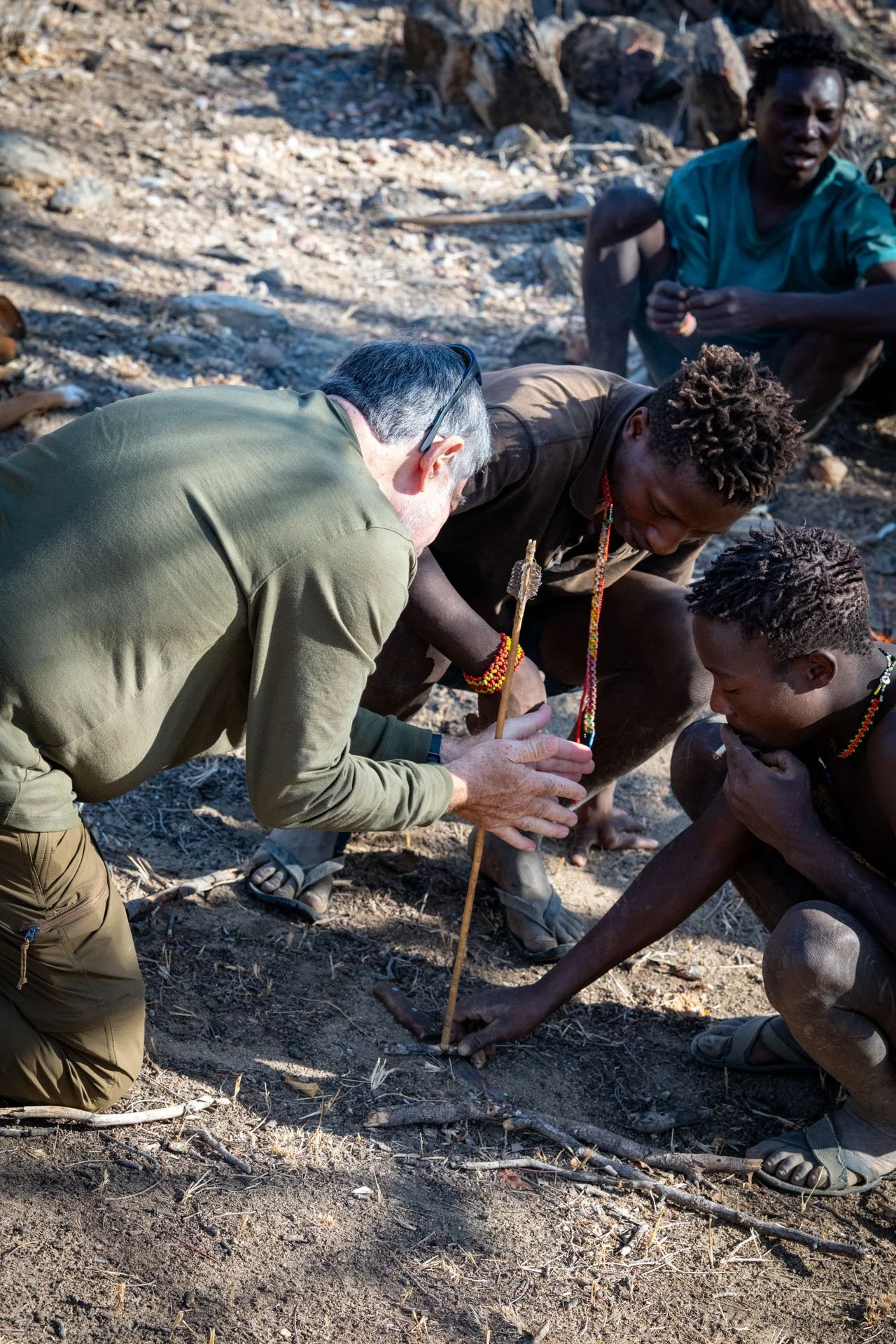 A man with gray hair and glasses kneels on the ground, helping two young men with dreadlocks dig a hole in the dirt with sticks, while another man in a teal shirt watches in the background.