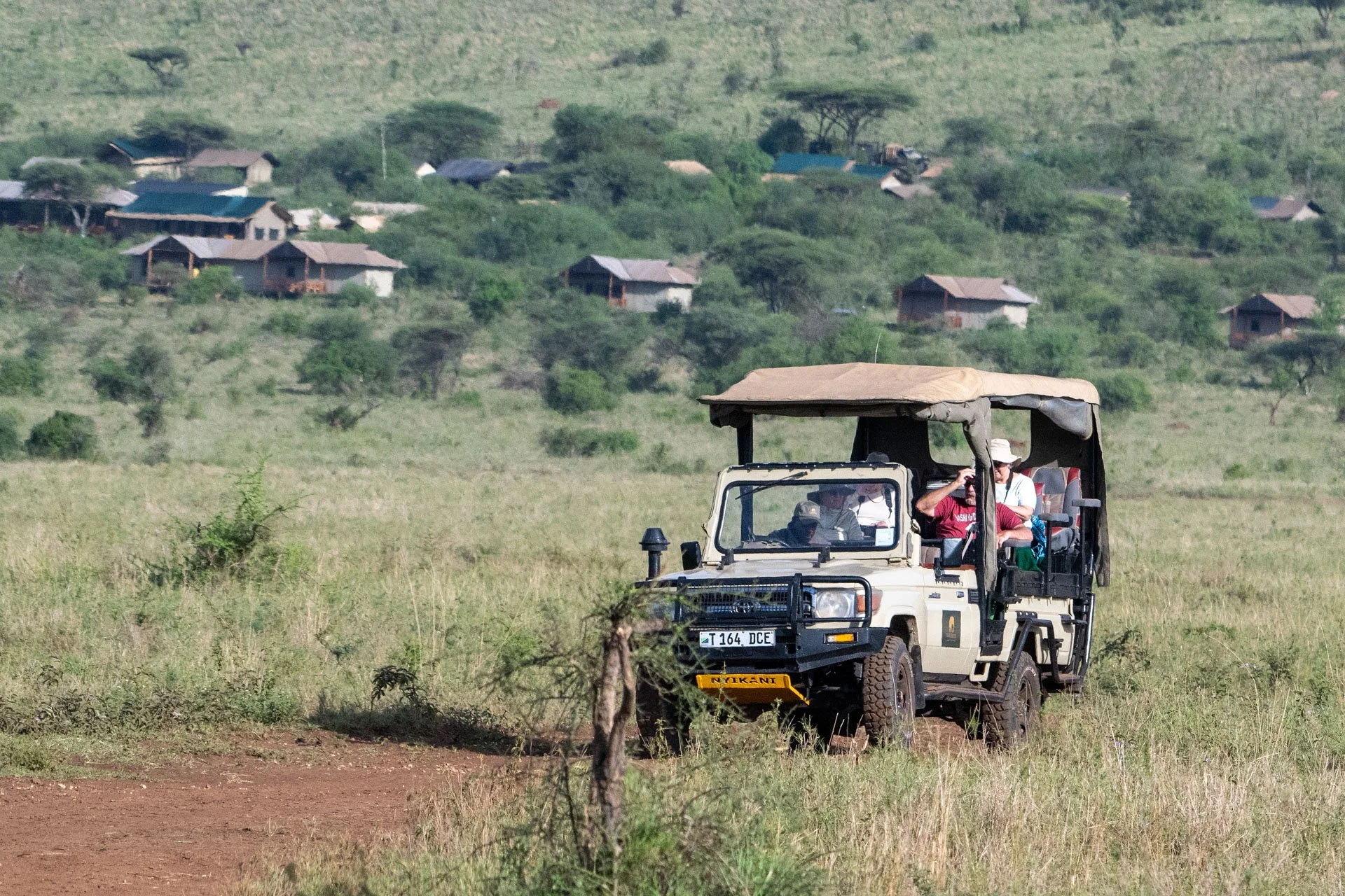 A safari vehicle with passengers on a dirt trail in a grassy landscape with scattered small bushes. In the background, there are hills with numerous small huts and houses.