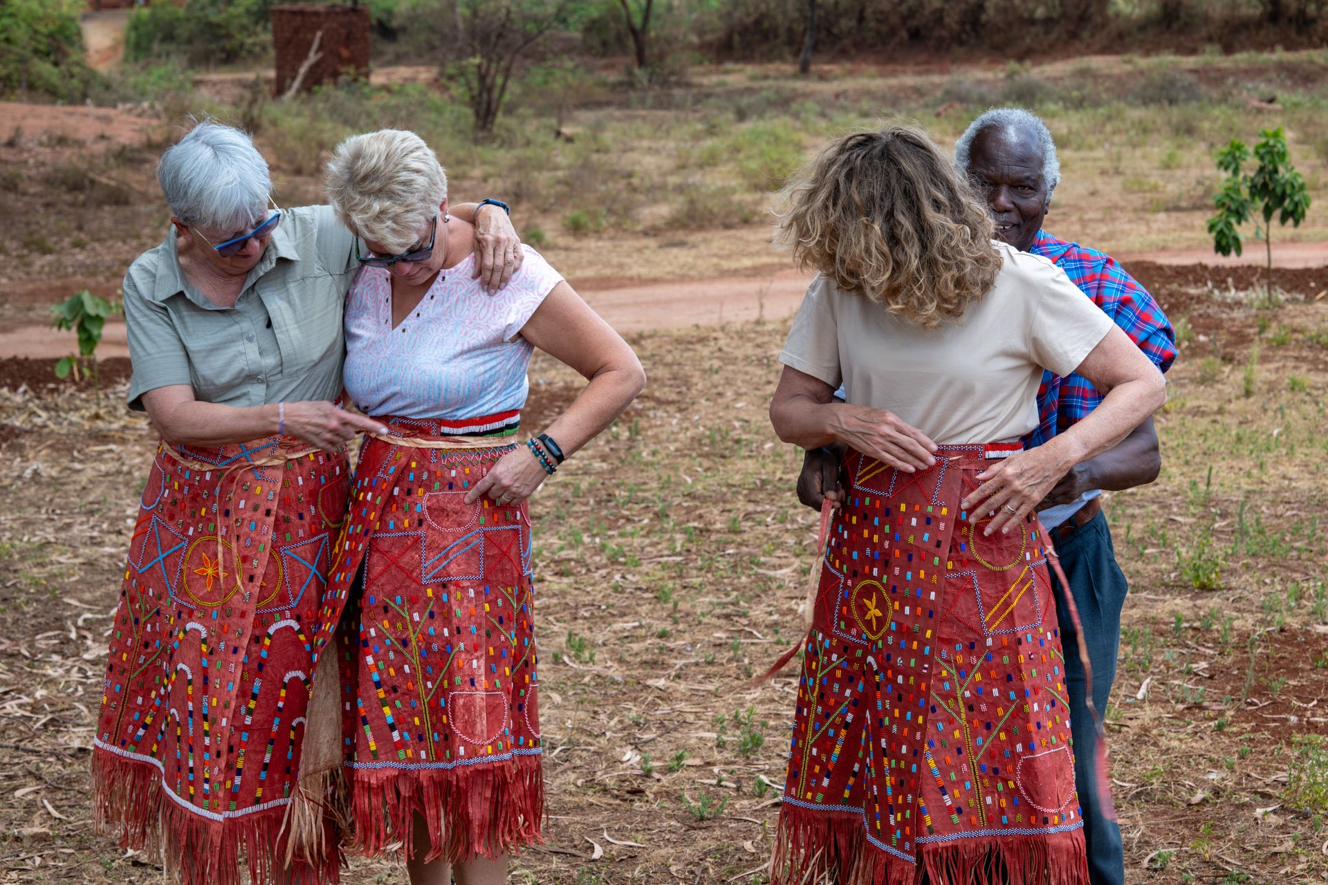 Four older adults wearing colorful, beaded skirts are gathered outdoors on a dirt field, with three women and one man embracing and smiling.