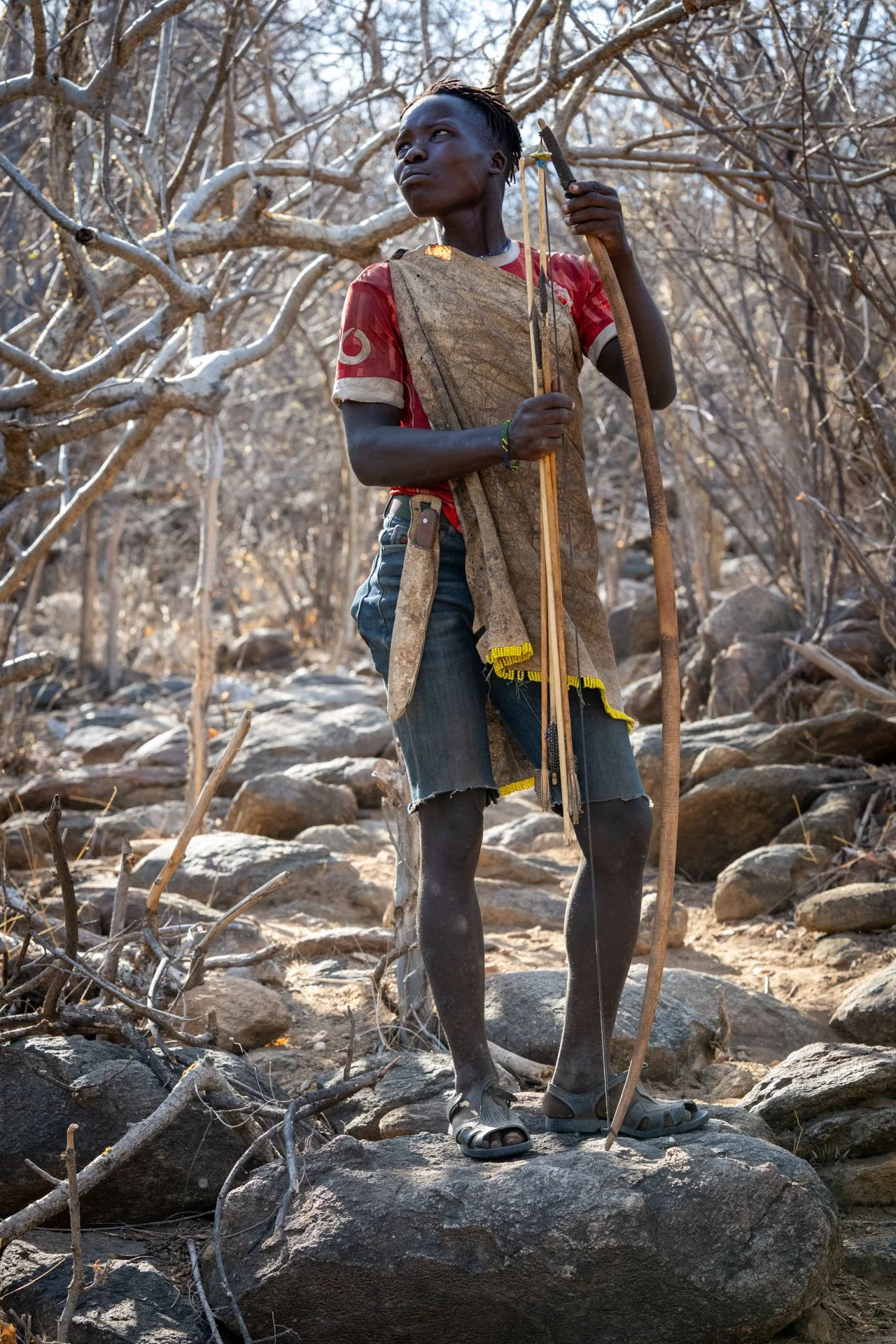 Young man standing on rocks in a dry, leafless forest holding a bow and arrows, wearing a long apron, ripped shorts, and slide sandals, gazing into the distance.