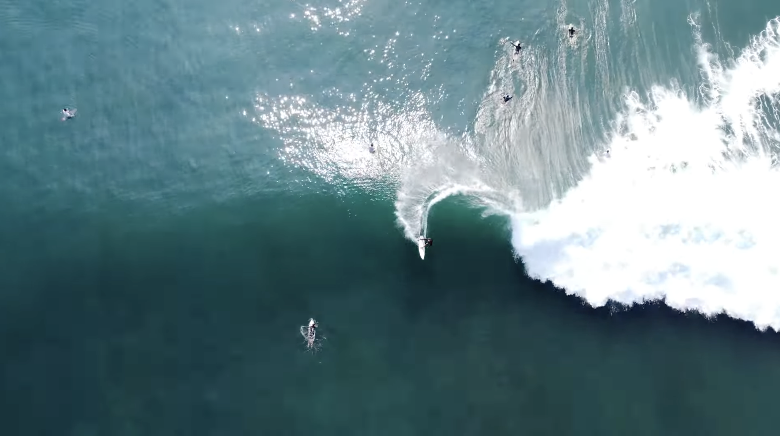 An aerial view of a surfer riding a large wave in the ocean with several other surfers in the water.