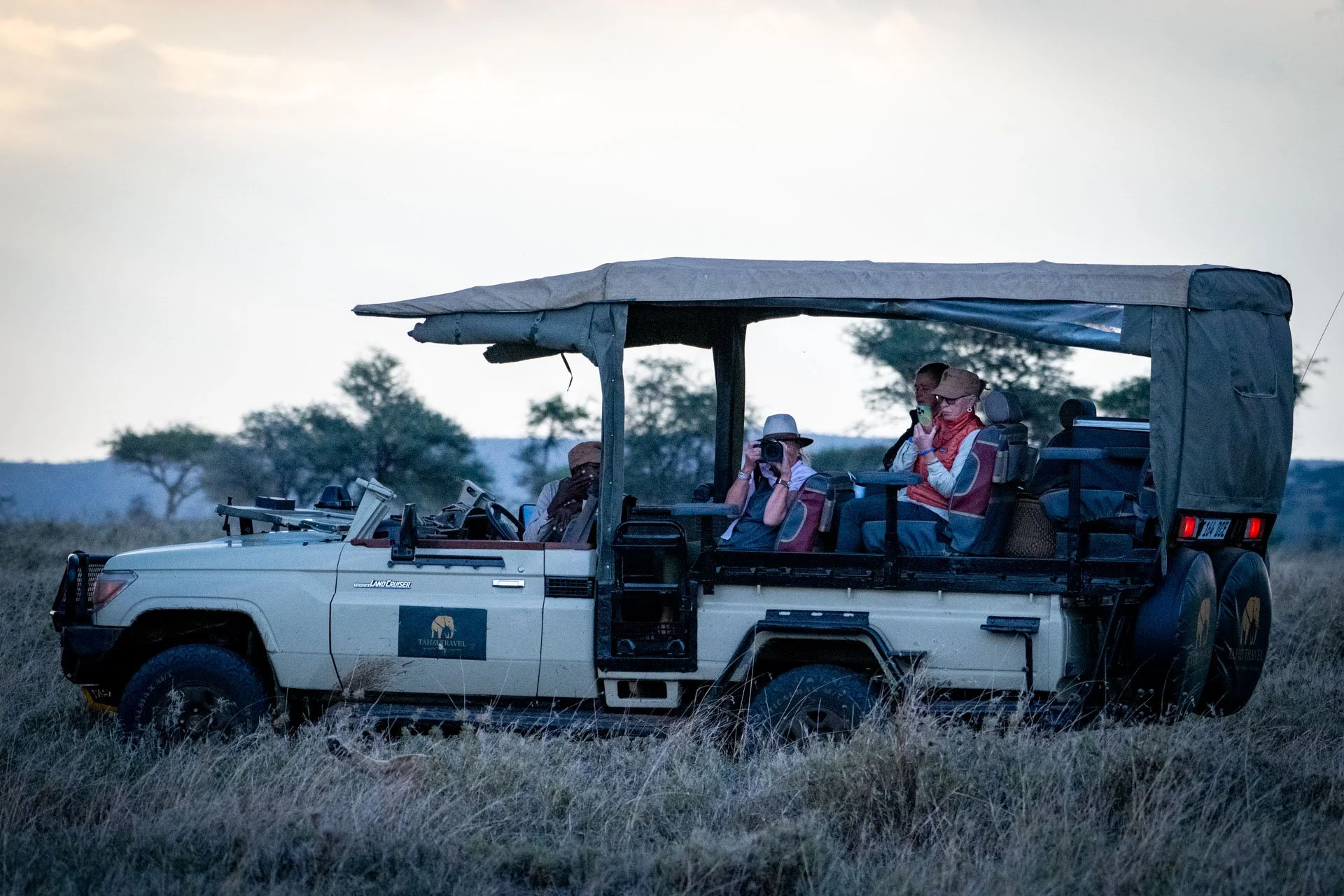 A group of tourists on an open safari vehicle in a grassy savannah, some taking photographs, during sunset or dusk with trees in the background.