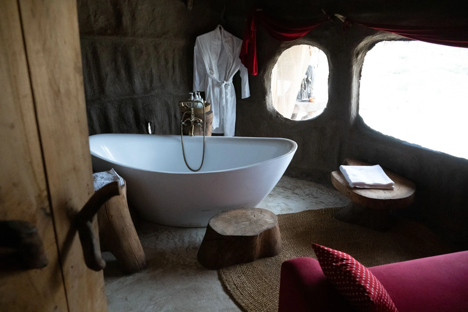 A rustic bathroom with a free-standing white bathtub, a wooden stool, a wooden bench with towels, a white bathrobe hanging, and two small windows in a dark stone wall.