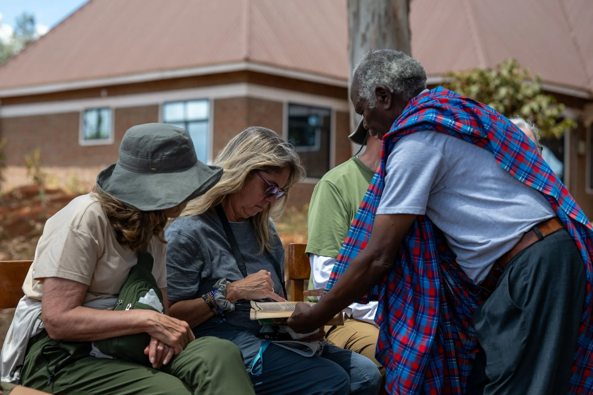 Group of diverse people sitting outdoors, looking at a book, with a man presenting or explaining something to them.