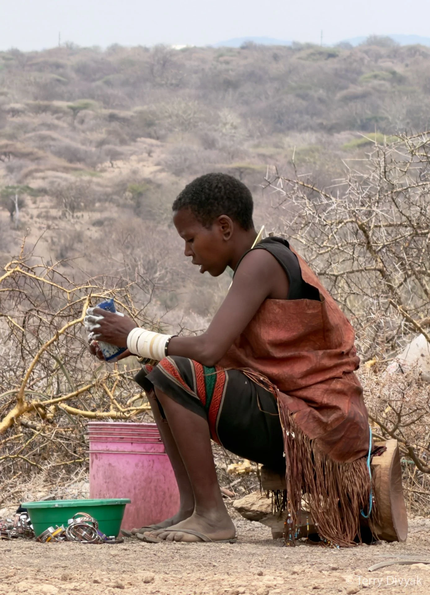 A person sitting outdoors in a dry, mountainous landscape, wearing traditional clothing and jewelry, holding an object in their hands, with various items in front of them.