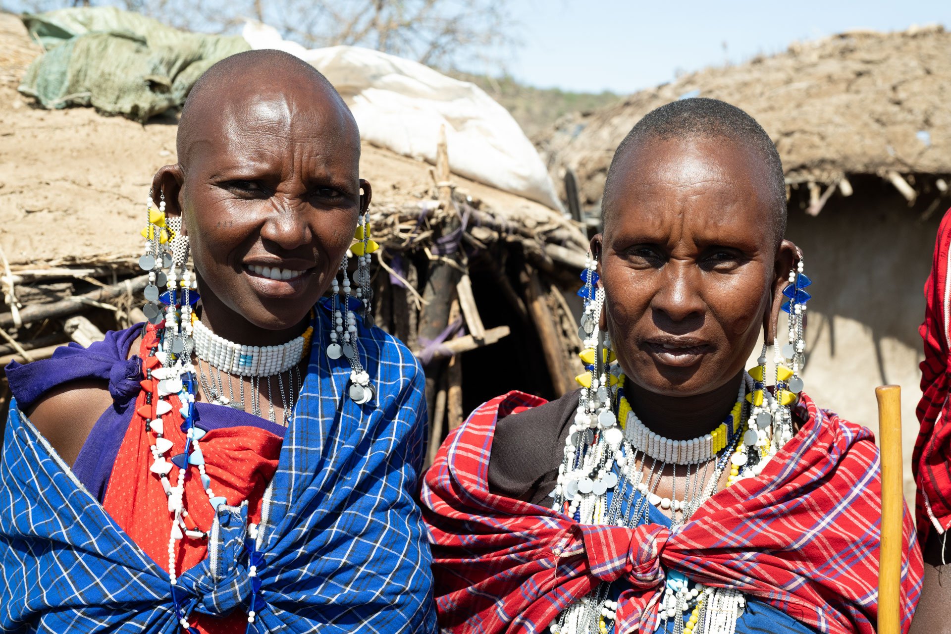 Two women from an African tribe dressed in colorful traditional clothing and elaborate beaded jewelry, standing outdoors with huts in the background.