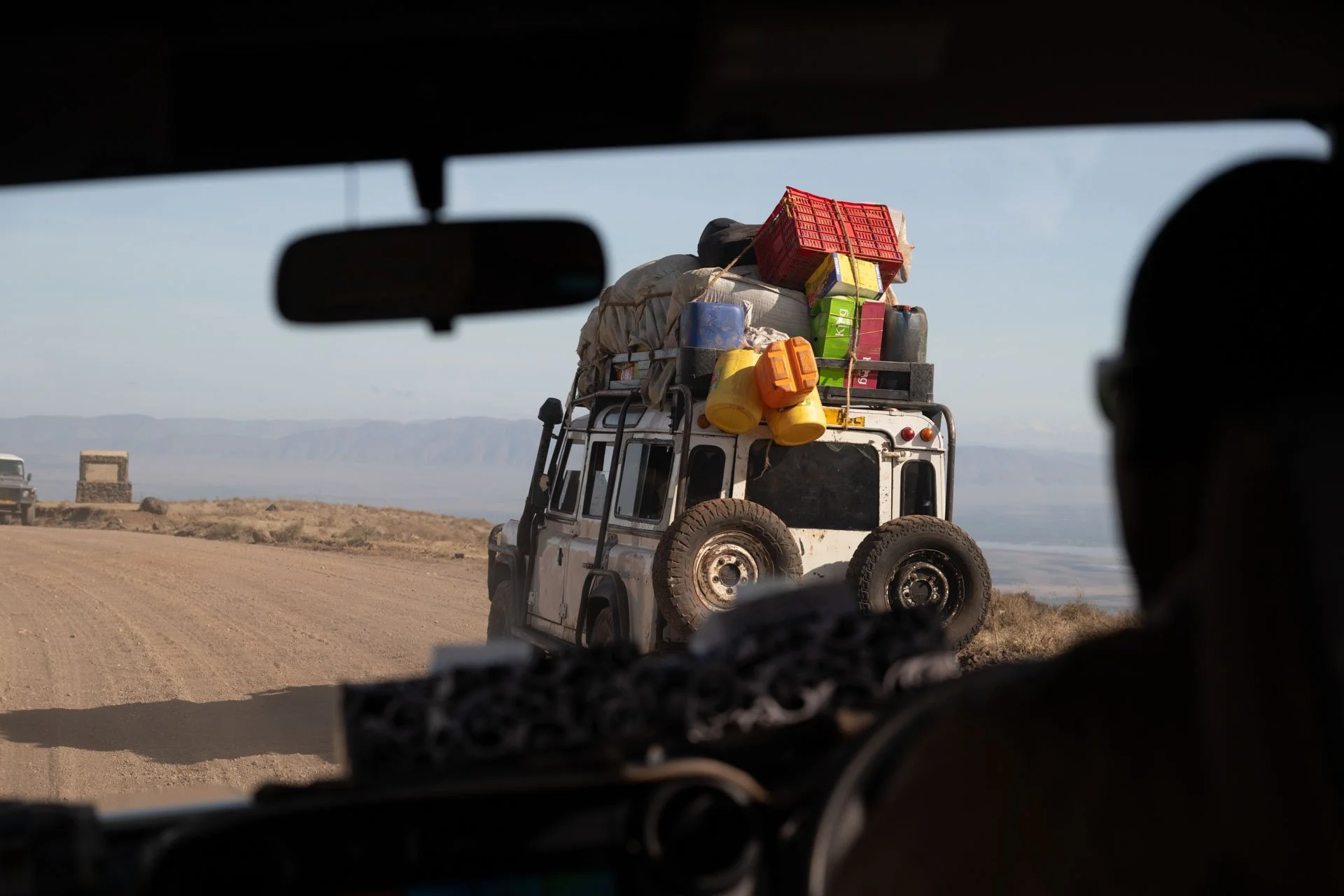 View from inside a vehicle looking at a loaded off-road vehicle with luggage and supplies on its roof, traveling on a dirt road in a desert landscape.