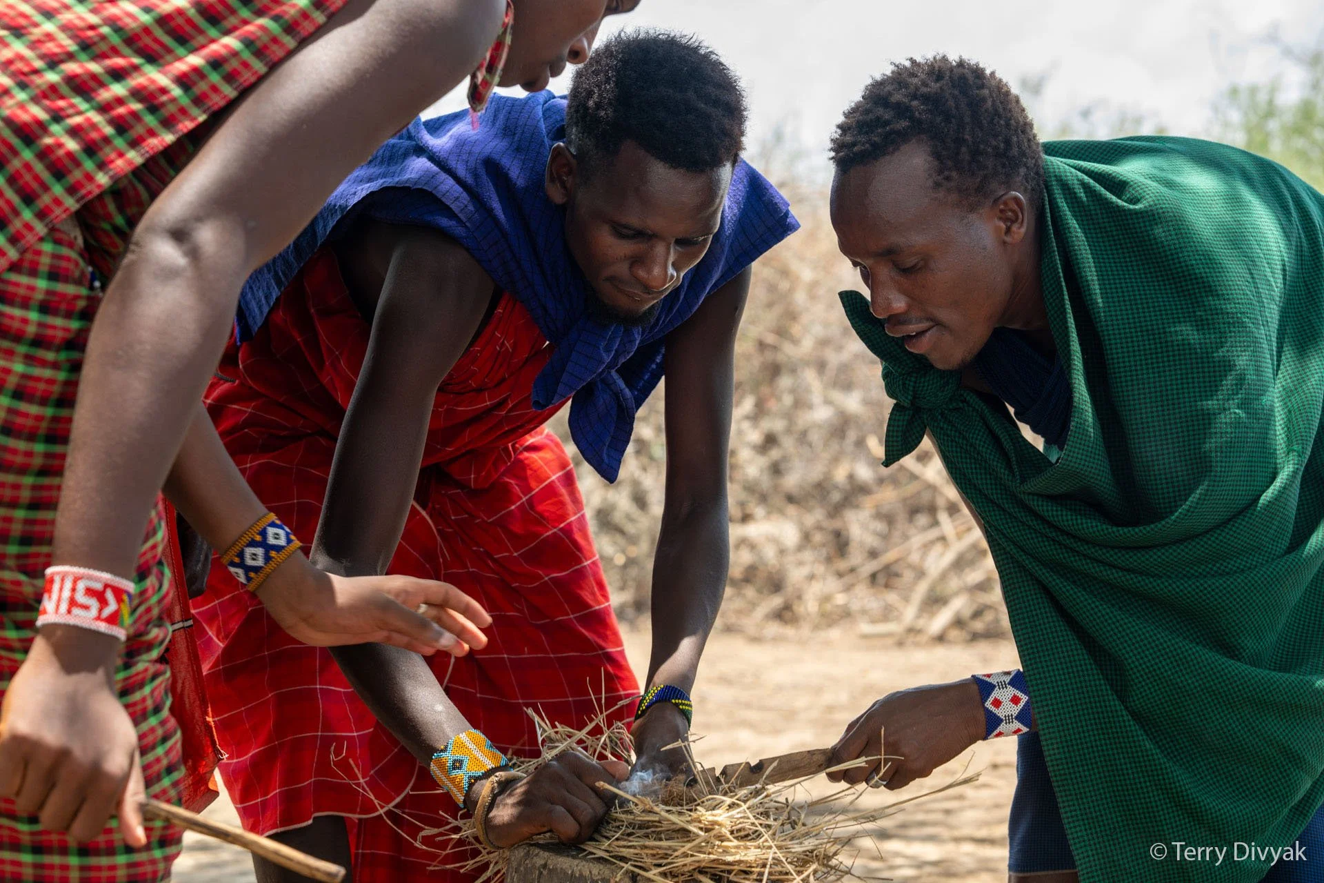 Two men from the Maasai tribe, dressed in traditional colorful clothing, are gathered around a small fire outdoors. They are using sticks to tenderly tend to the fire.