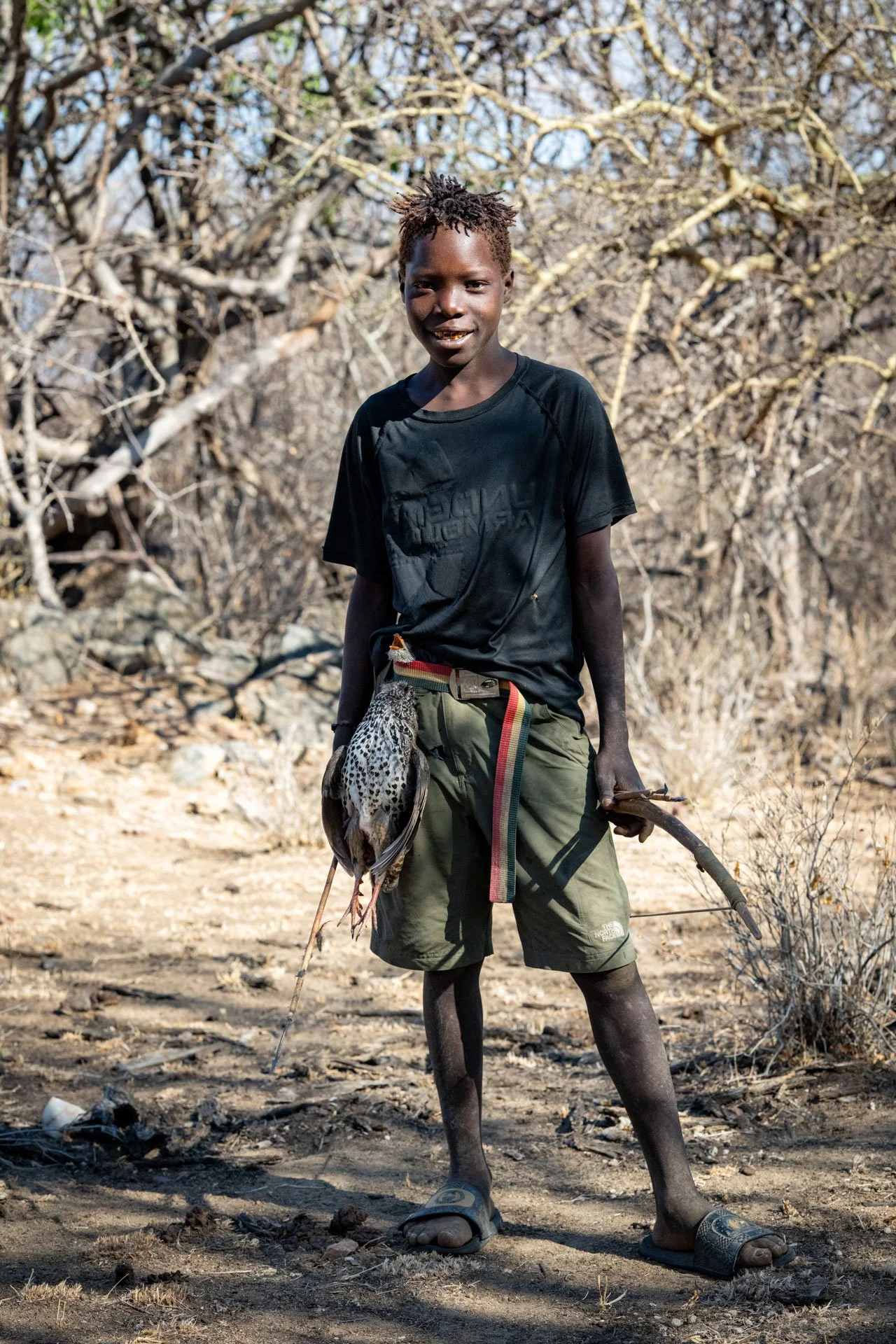 Young boy standing outdoors on dry land with leafless trees behind him, holding a bird of prey.