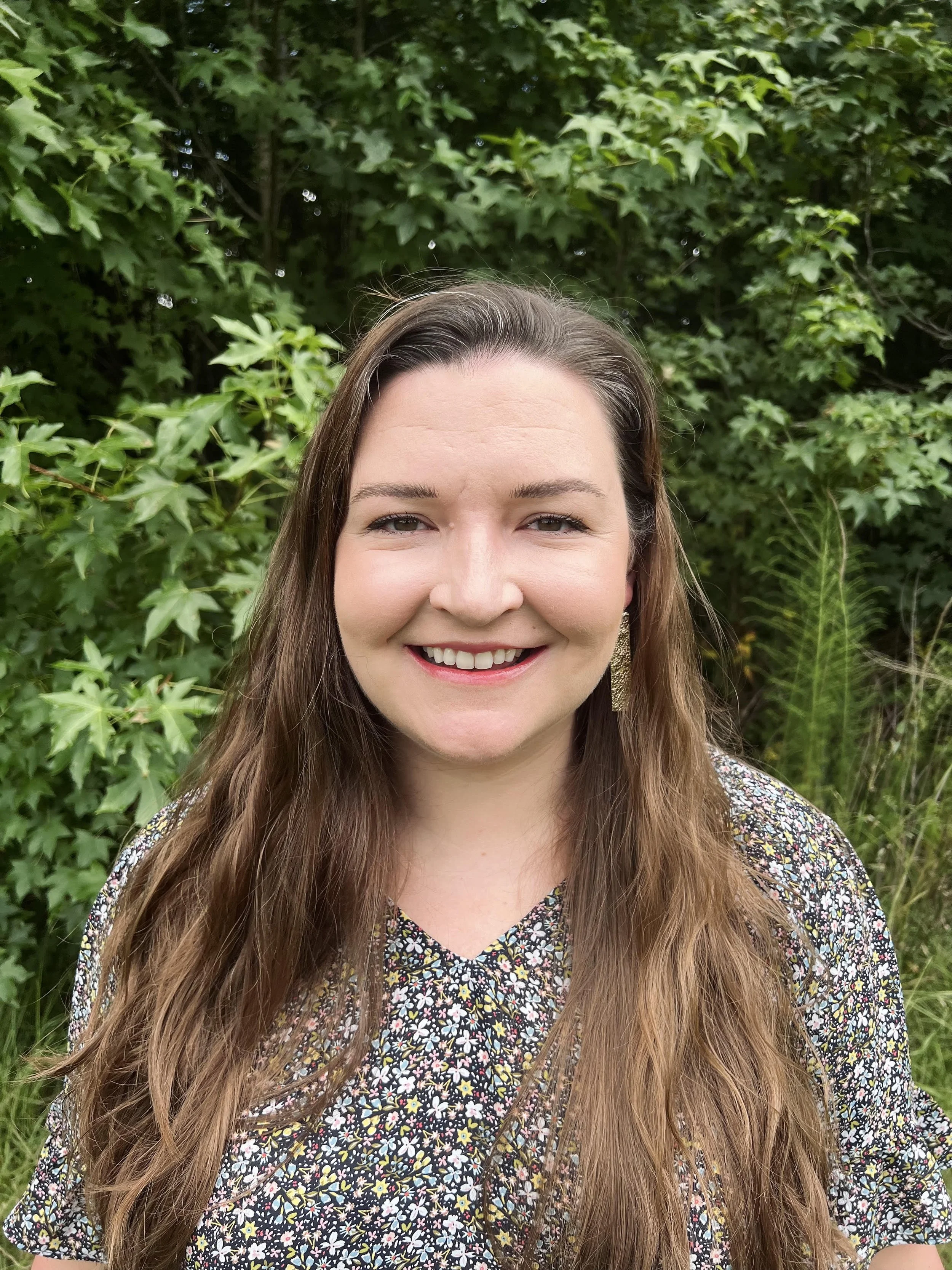 A smiling woman with long brown hair, wearing a floral blouse, standing outdoors in front of green foliage.