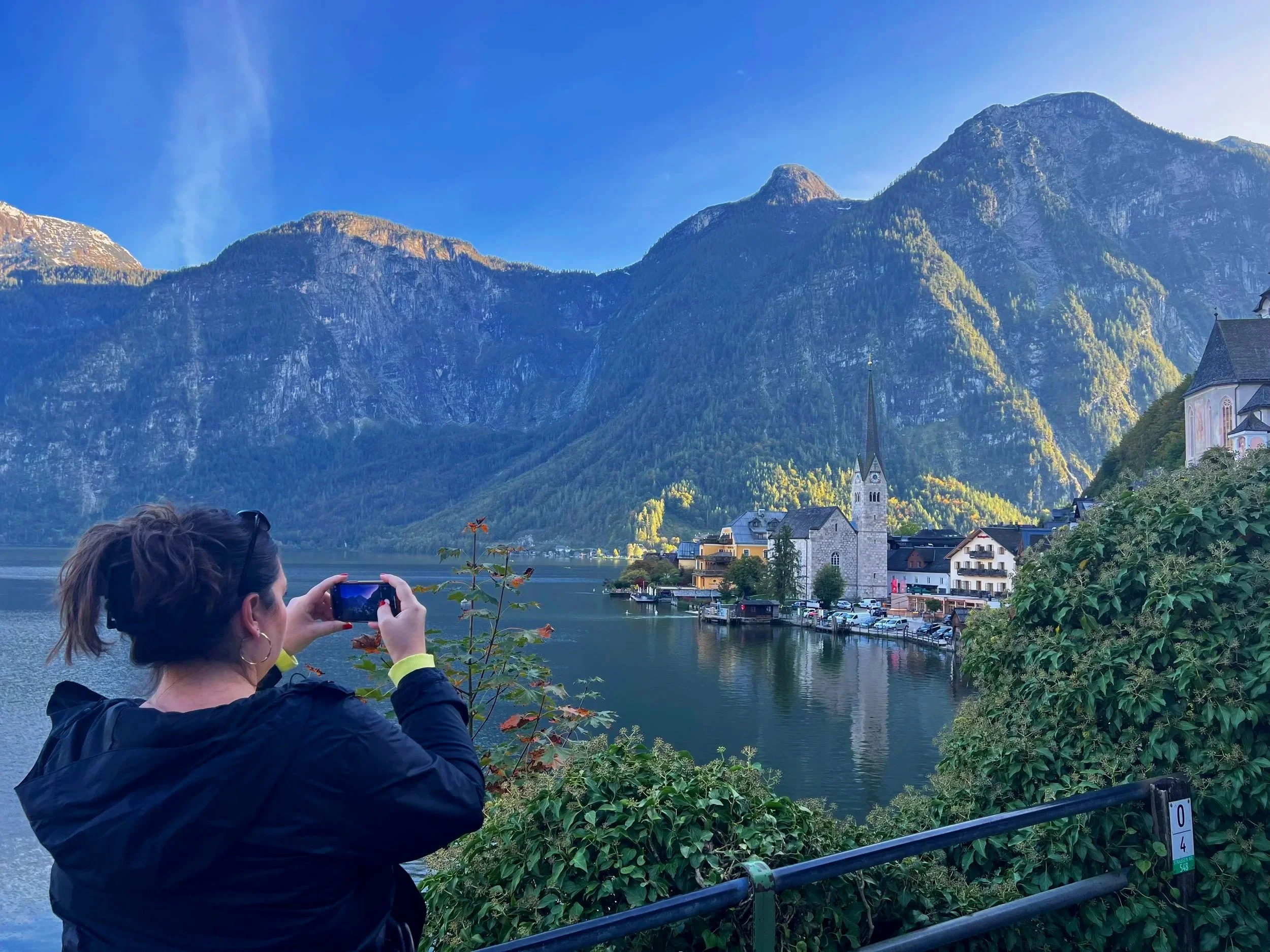 Woman with dark hair and sunglasses taking a photo of the scenic lakeside village with mountains in the background.