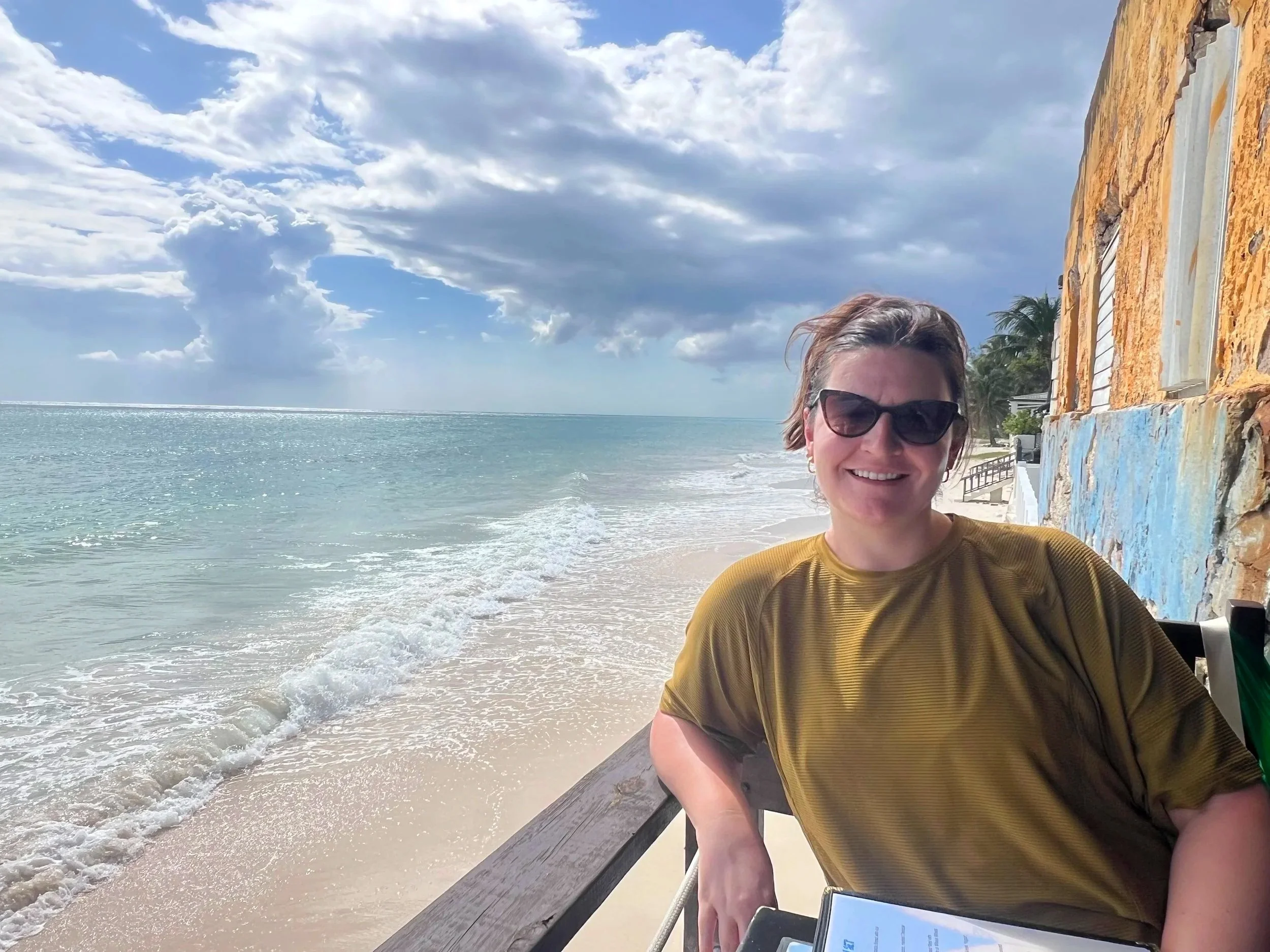 Smiling woman wearing sunglasses and a yellow shirt sitting on a seaside bench next to a colorful stone wall, with a beach, ocean, and partly cloudy sky in the background.
