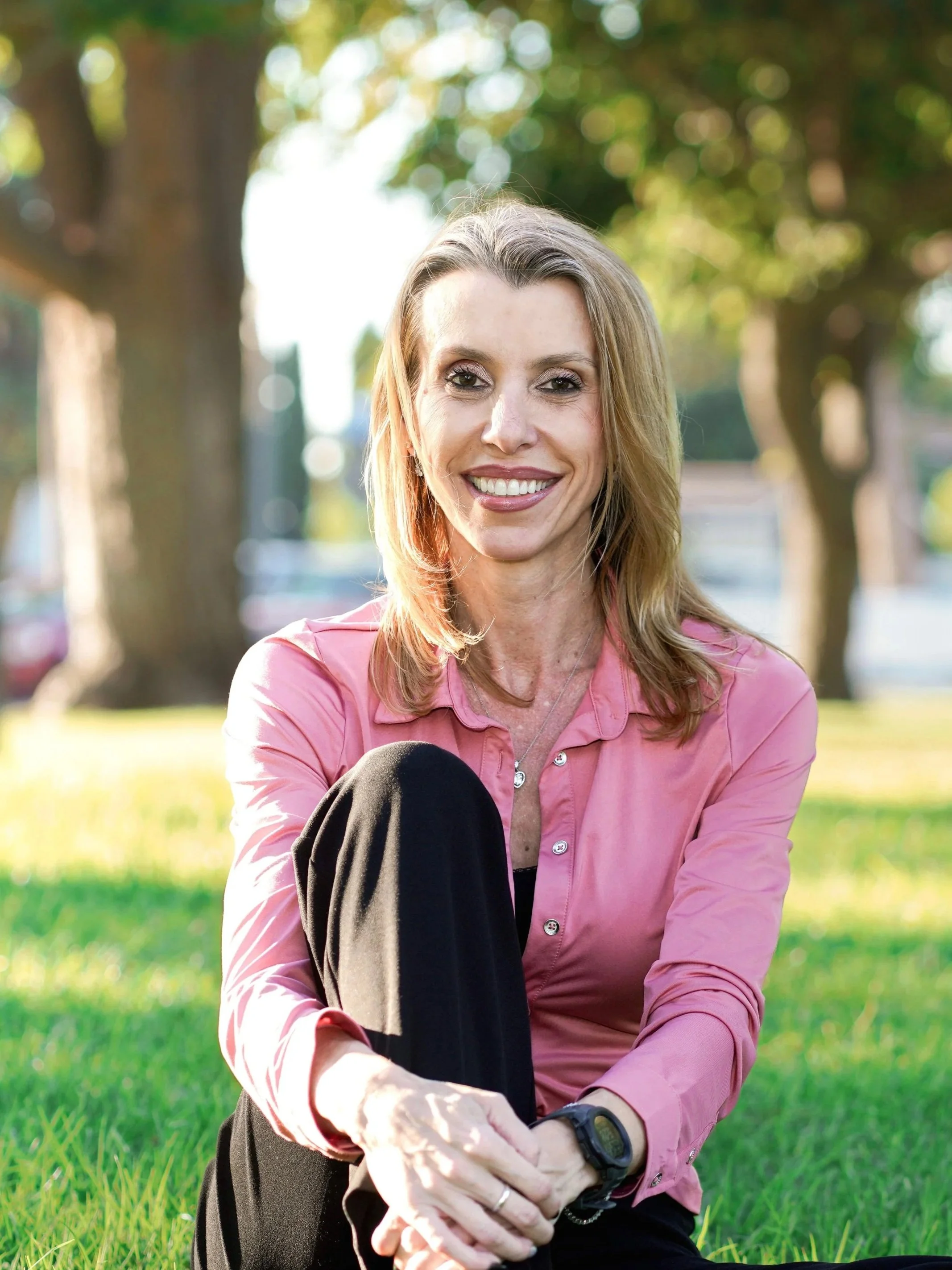 A woman sitting on grass in a park, smiling and wearing a pink shirt and black pants, with trees and sunlight in the background.