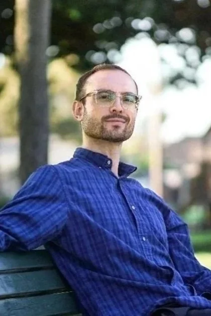 A man with glasses and a beard sitting on a park bench, wearing a blue button-up shirt, with trees and a blurred background.