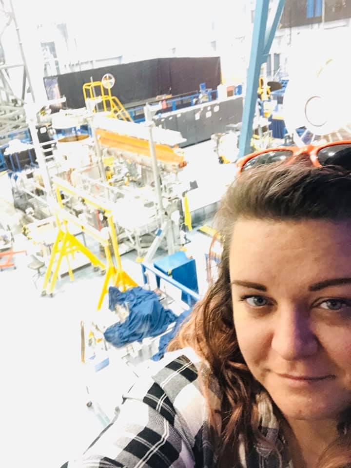 Selfie of a woman with brown curly hair and sunglasses on her head, standing in a factory or industrial setting with complex machinery and equipment behind her.
