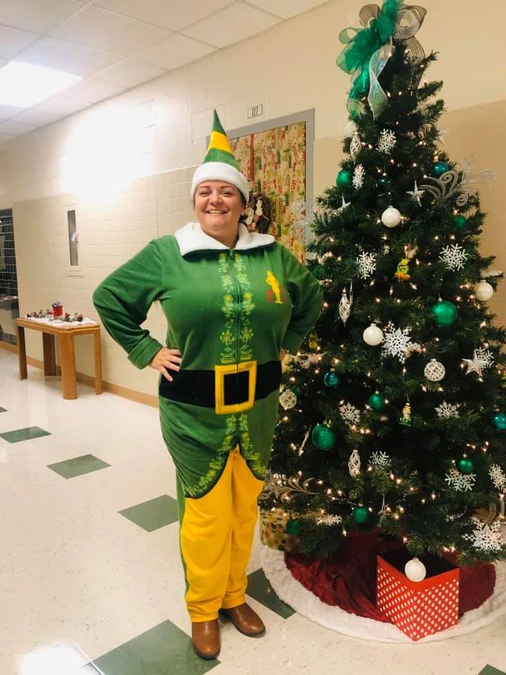 A woman dressed as an elf standing next to a decorated Christmas tree in a hallway.