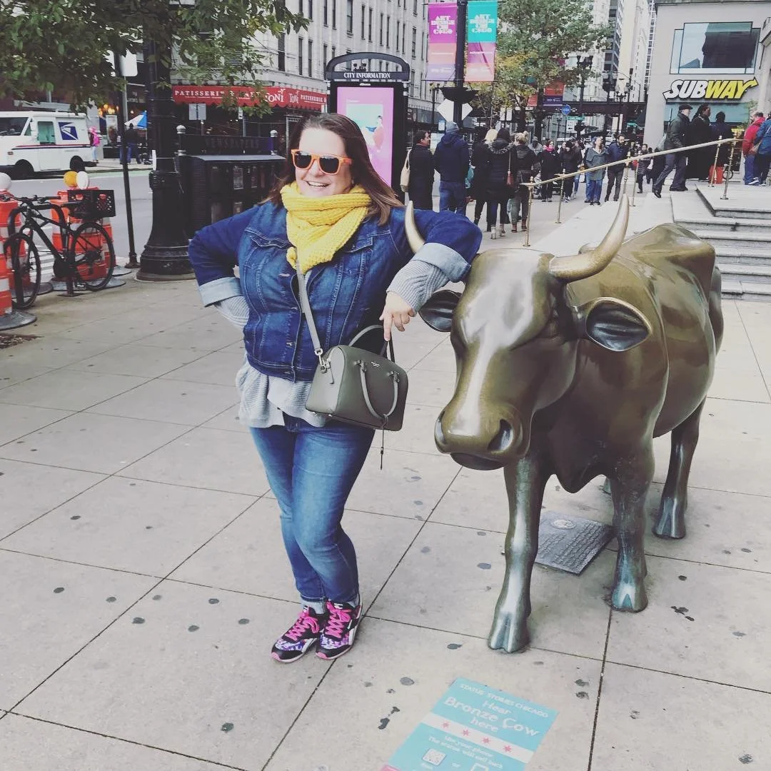 A woman with sunglasses, a yellow scarf, denim jacket, and jeans stands next to a bronze statue of a bull on a city sidewalk. She is smiling and has her hand on the bull statue's head.