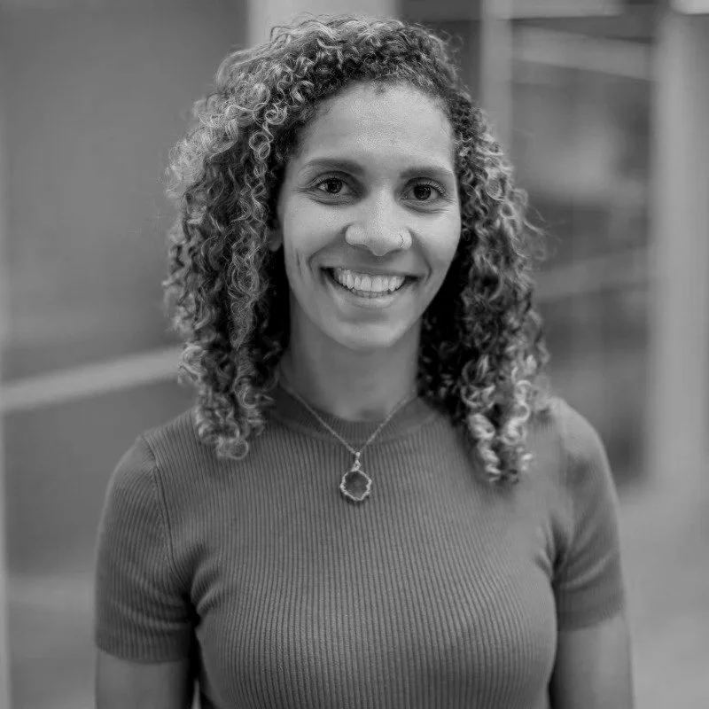 A man with curly dark hair, clear glasses, a beard, and a nose ring, wearing a dark green sweater, sitting indoors with a plant visible in the background.