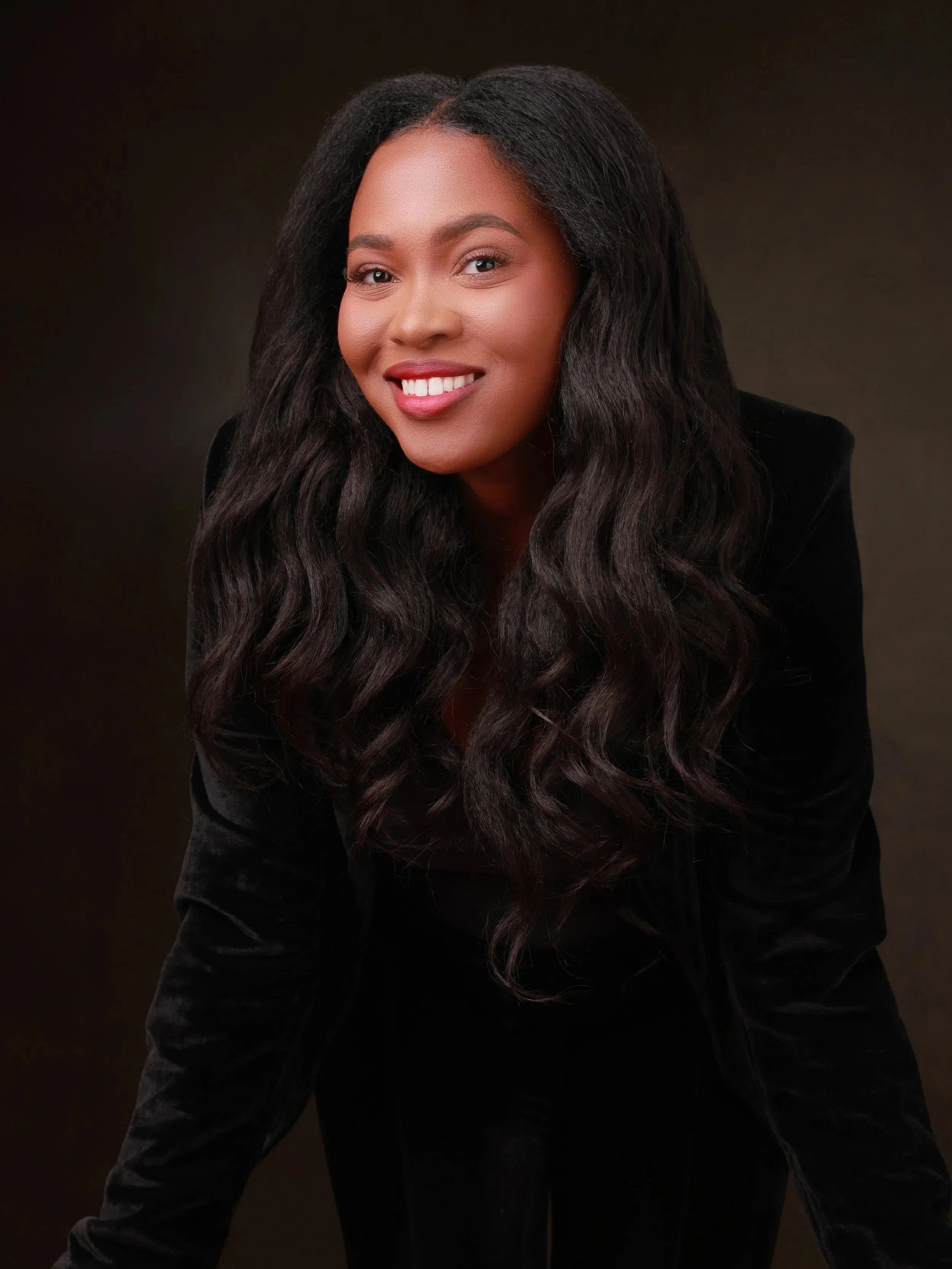 A young woman with long, wavy black hair and a bright smile, wearing a black velvet blazer, posed against a dark background.